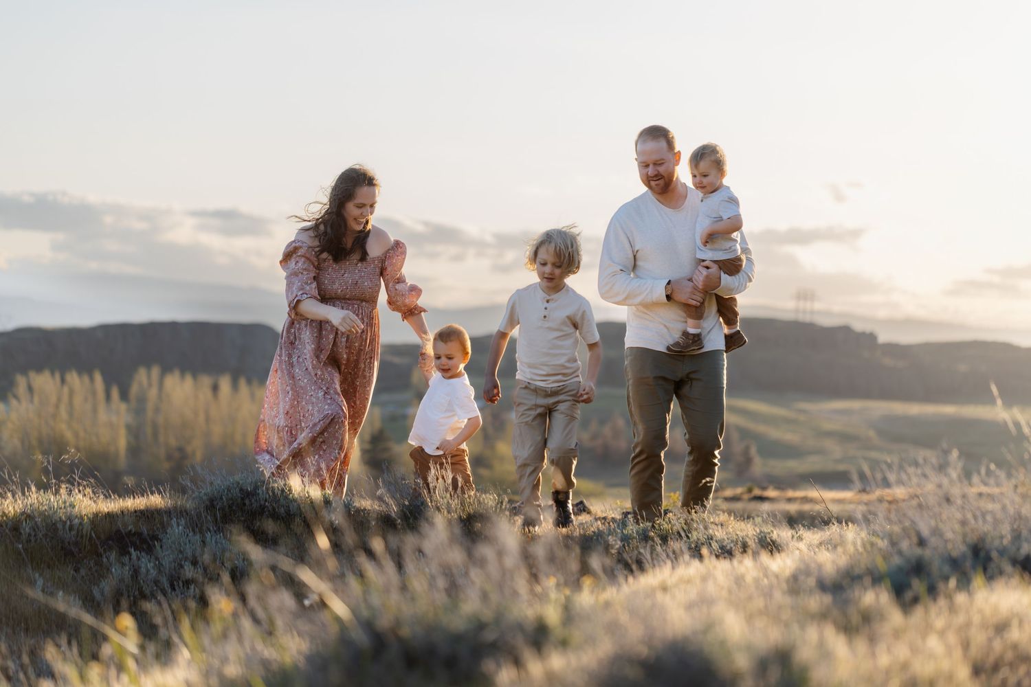 A family walks together through a sunlit meadow during golden hour as the sun sets behind rolling hills.