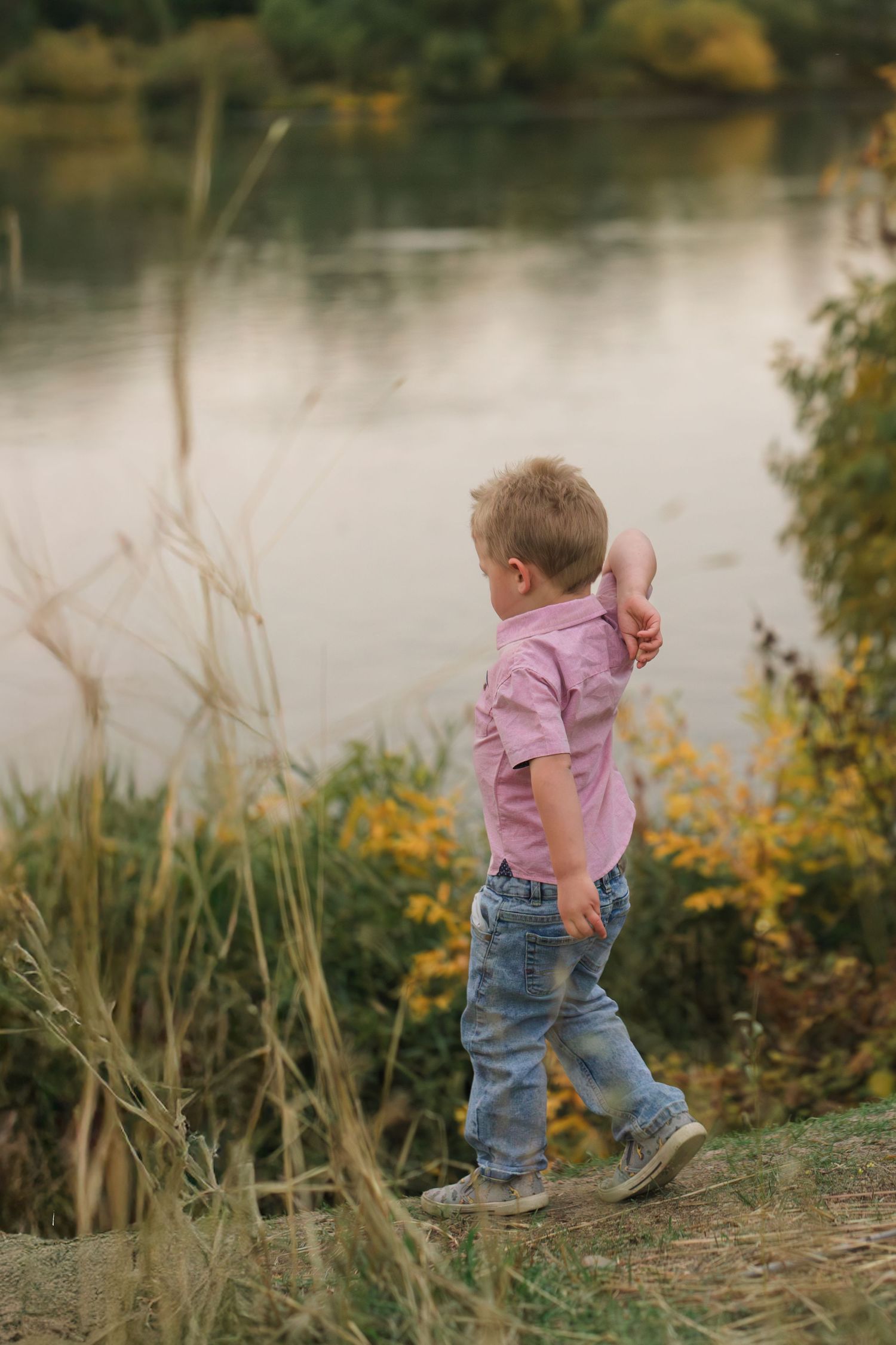 Small child in pink shirt and jeans walks along grassy riverbank with autumn foliage.