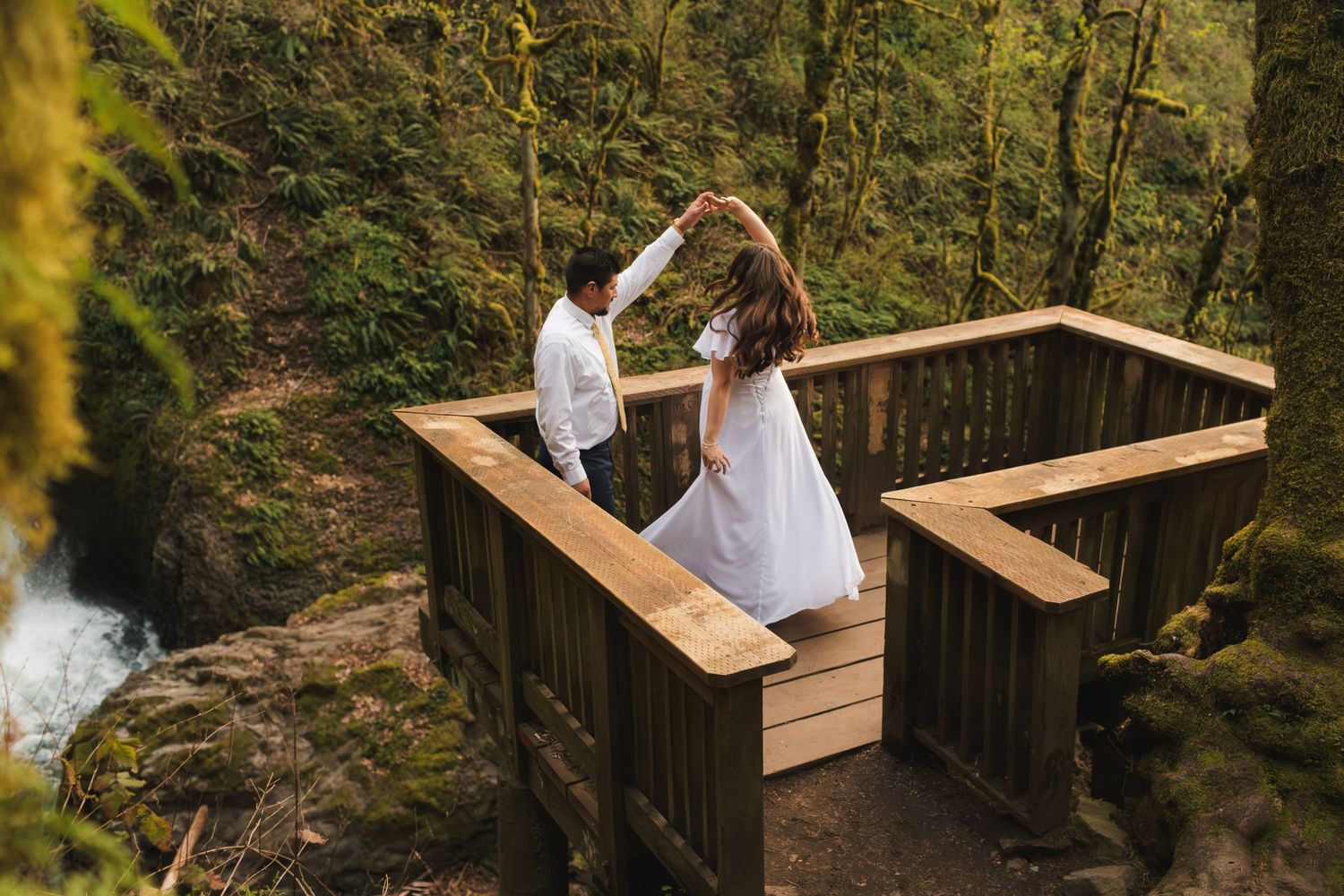 Couple dancing on a wooden overlook platform surrounded by lush green forest and waterfall in background.