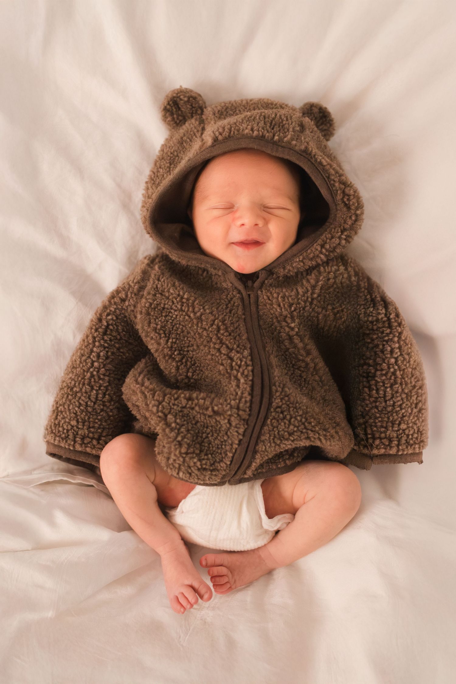 Smiling newborn baby wearing a cute brown teddy bear hoodie jacket lying on a white blanket.