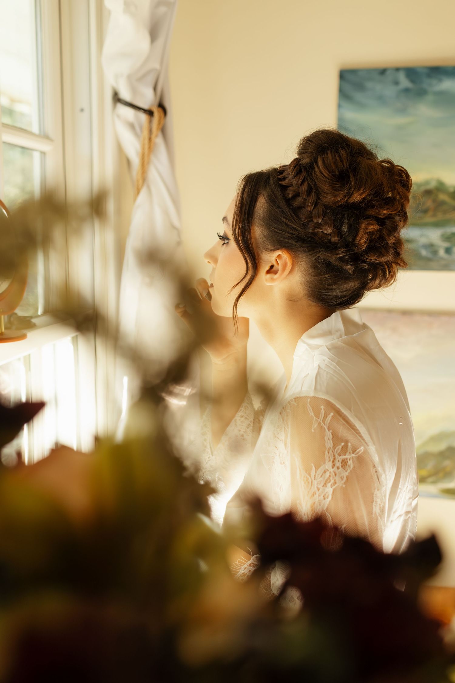 Soft morning light streams through window as bride gets ready in white silk robe.