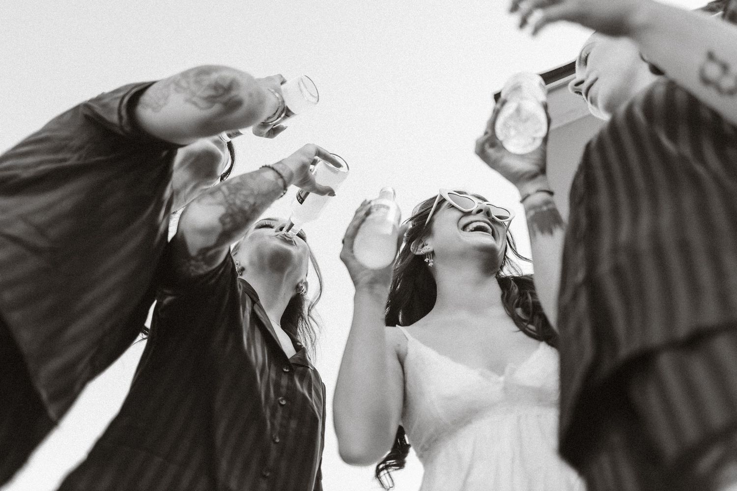 Black and white photo of bride and friends toasting with drinks at a wedding celebration outdoors.