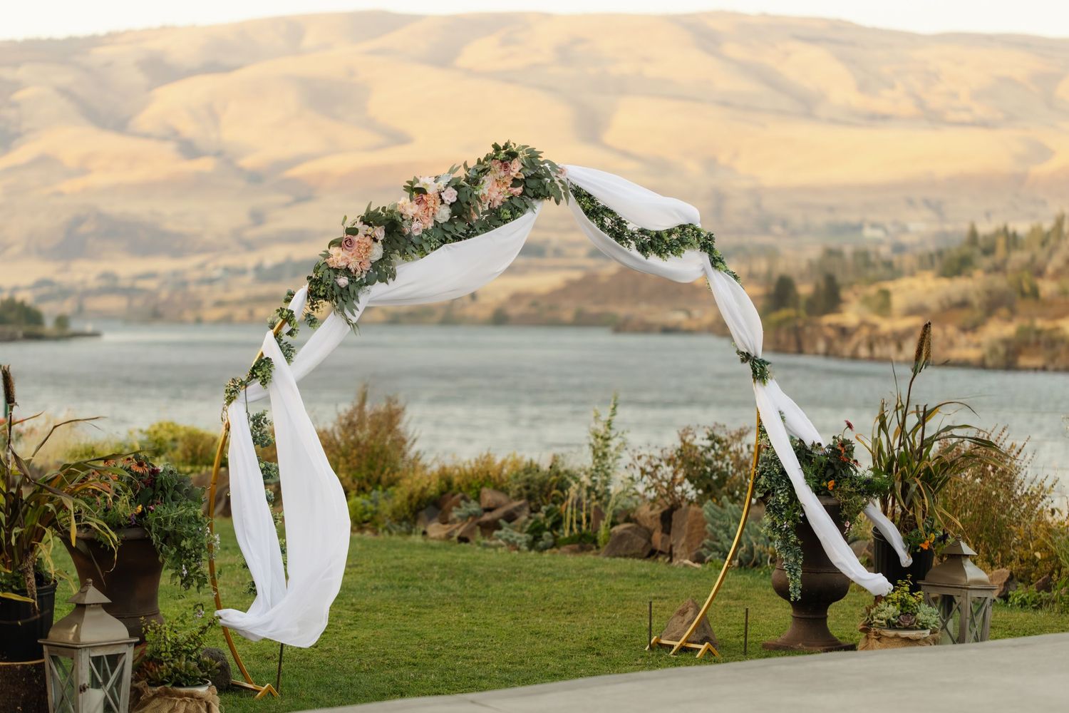 Floral wedding arch with white draping overlooking a scenic lake and golden hills backdrop.