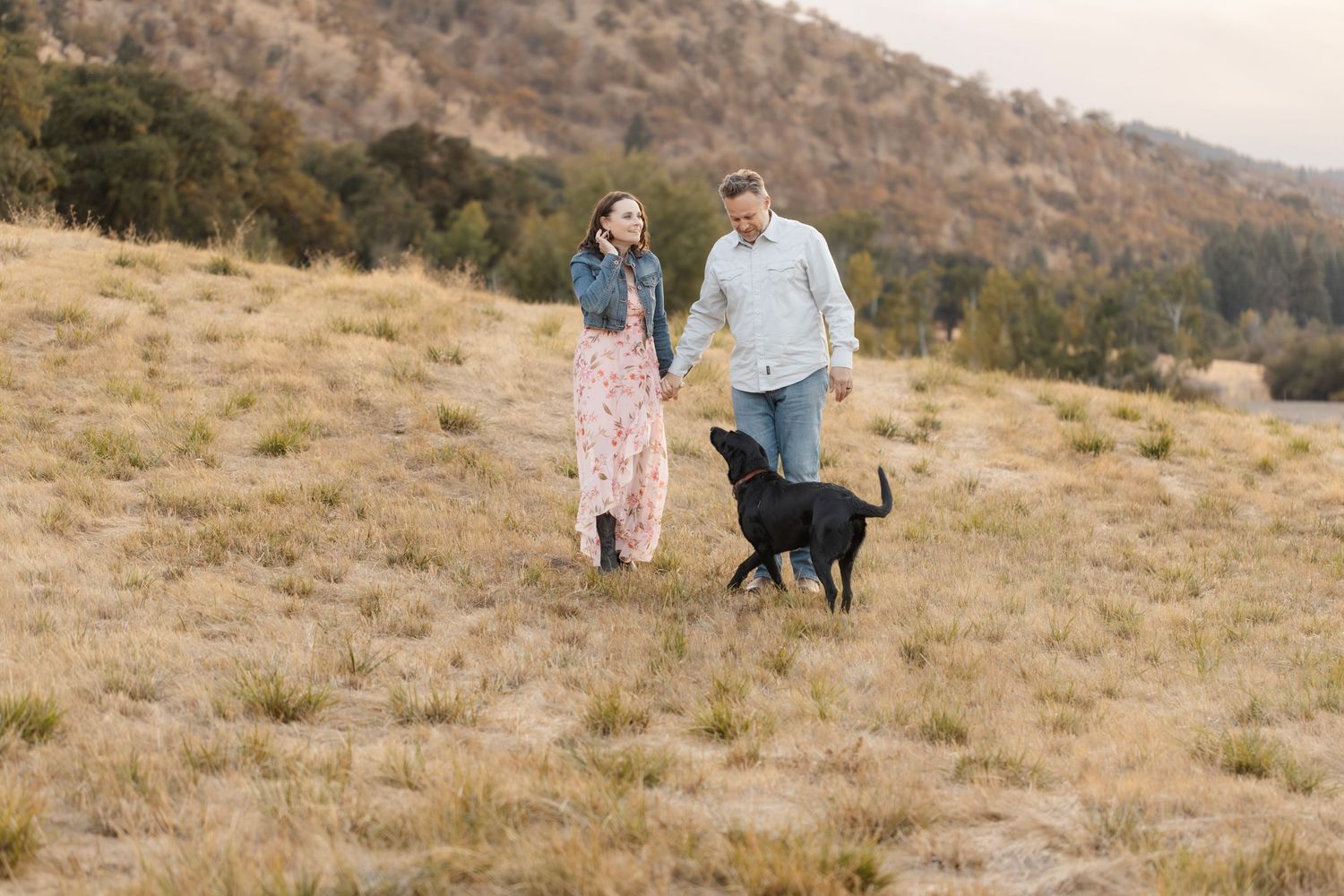 A couple and their dog enjoy a playful moment on a scenic hillside during golden hour.
