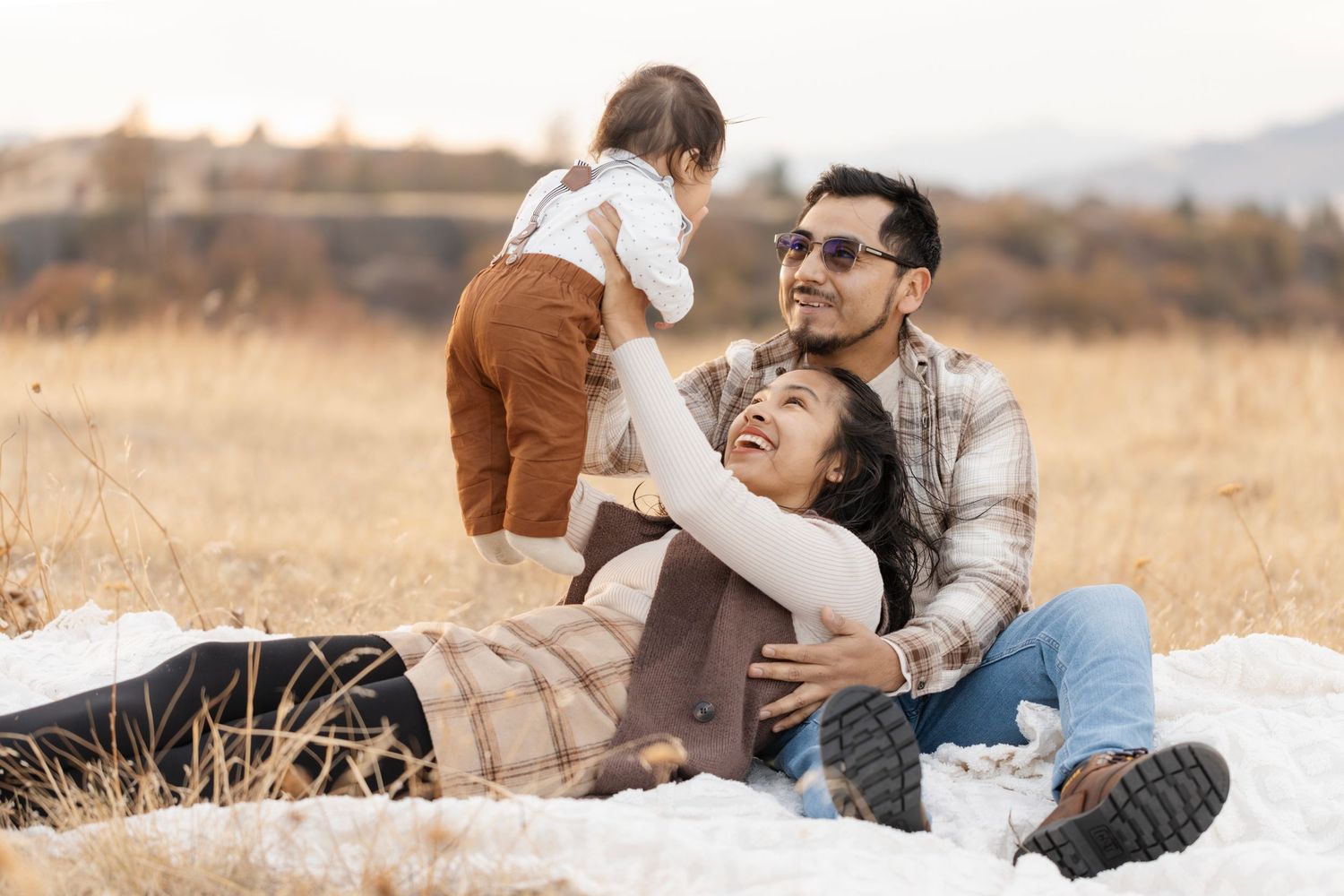 Playful family moment captured on blanket in golden field during sunset.