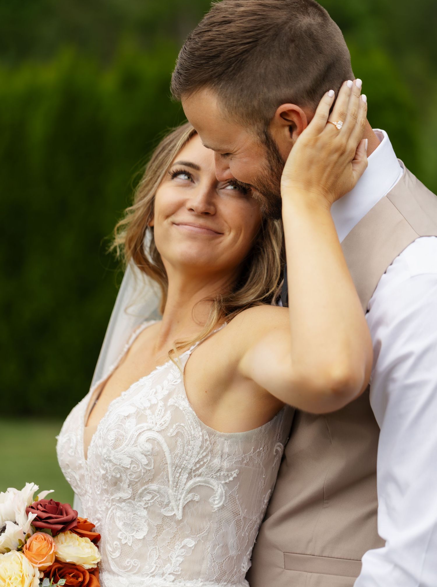A romantic wedding couple shares an intimate moment outdoors with lush greenery in the background.