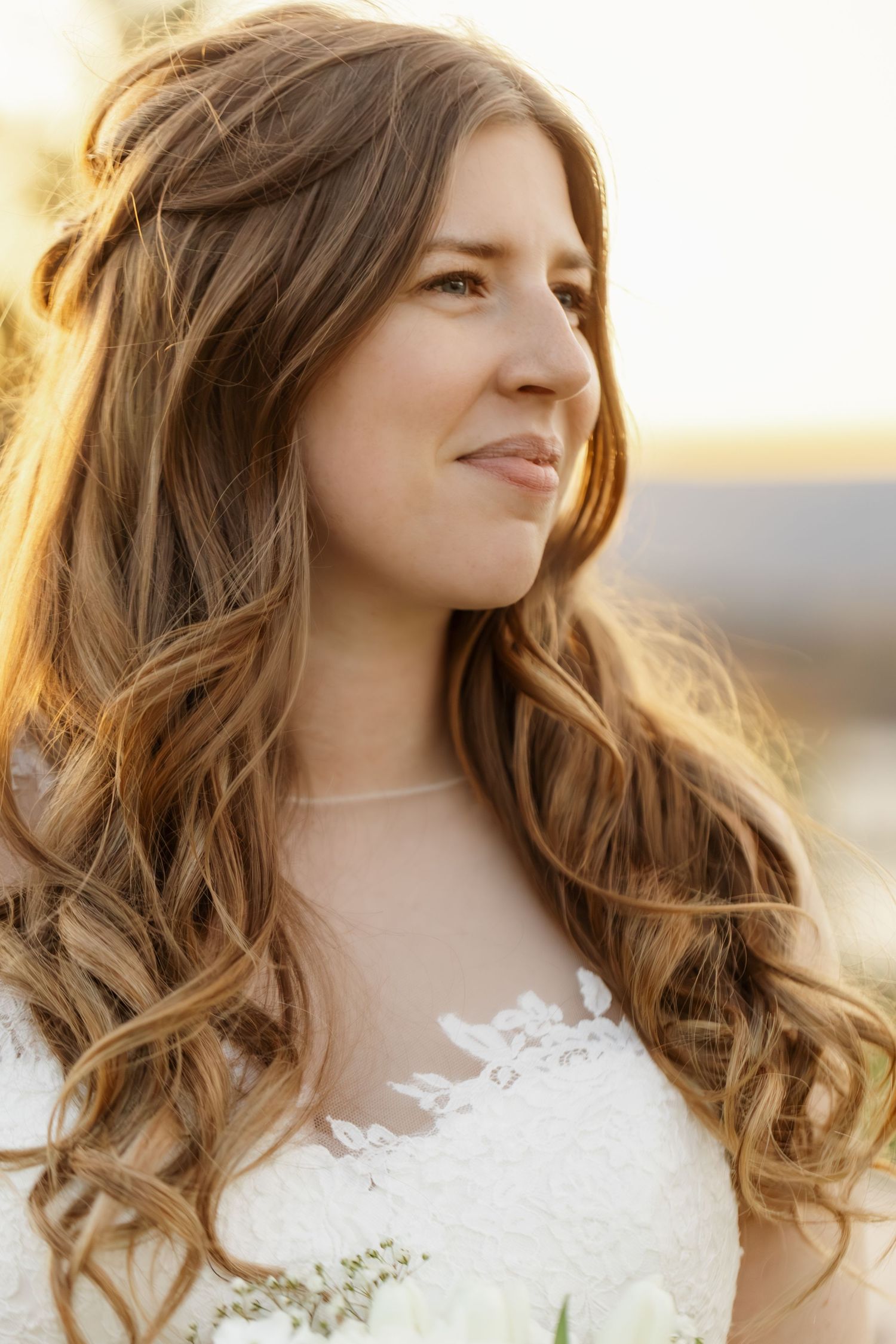 A bride in a white lace wedding dress with long wavy brown hair smiles during golden hour sunset.