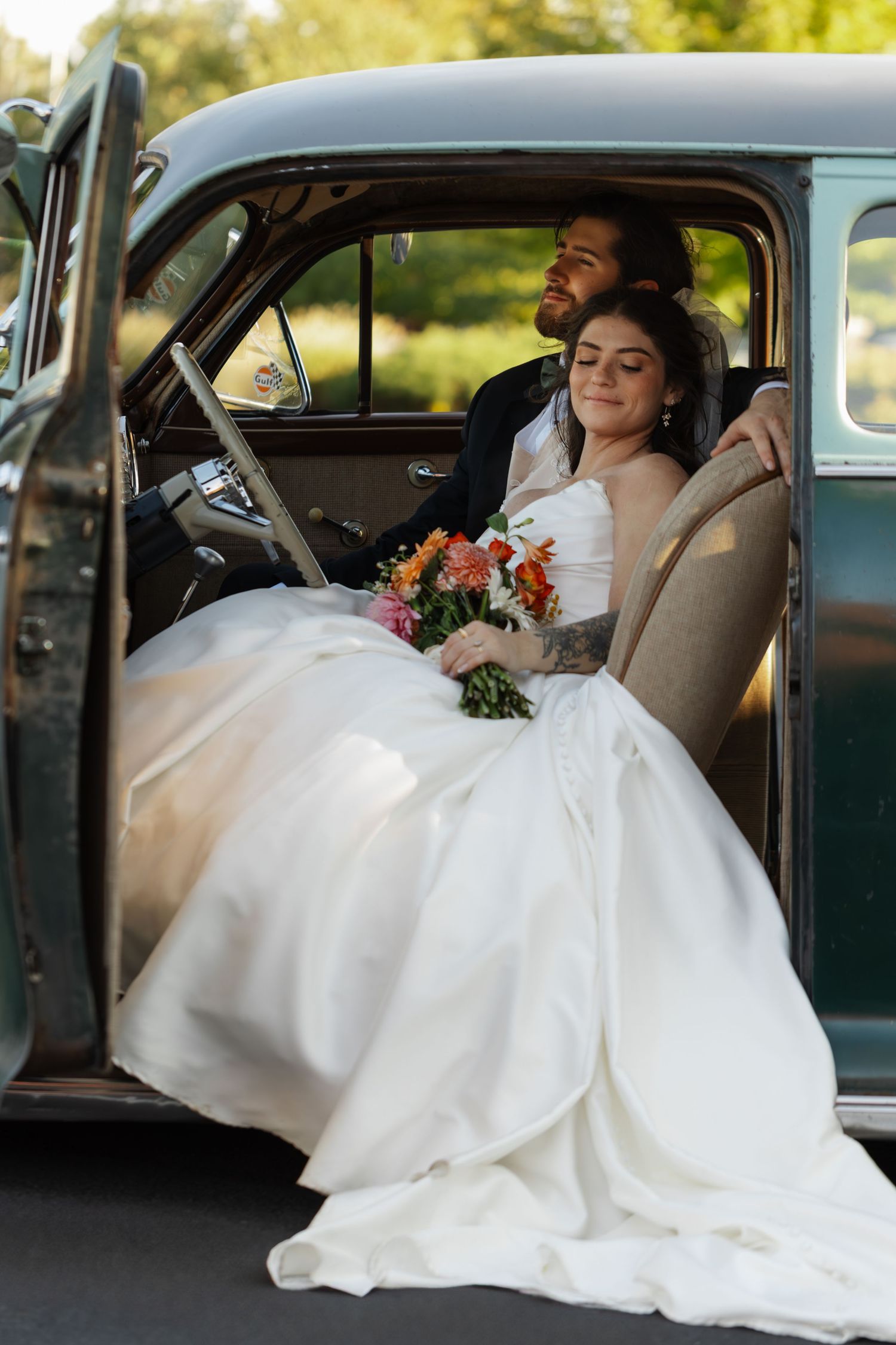 Wedding couple relaxing in the back seat of a vintage car with flowing white wedding dress.