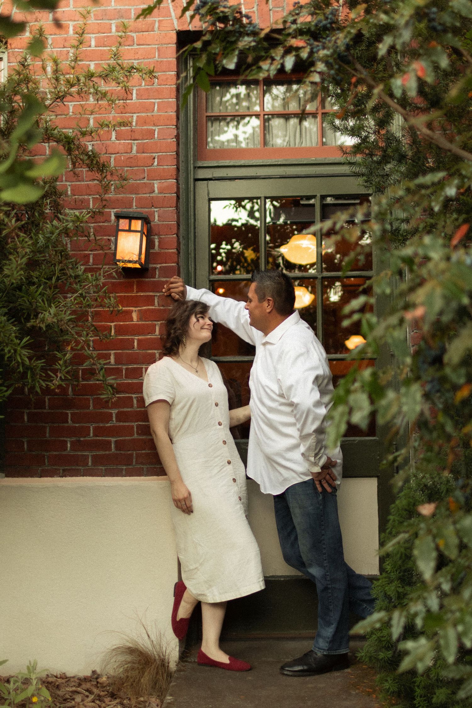 Couple posing by a red brick building with a glowing wall lantern, the man gently holding the hand of a woman in a white dress.