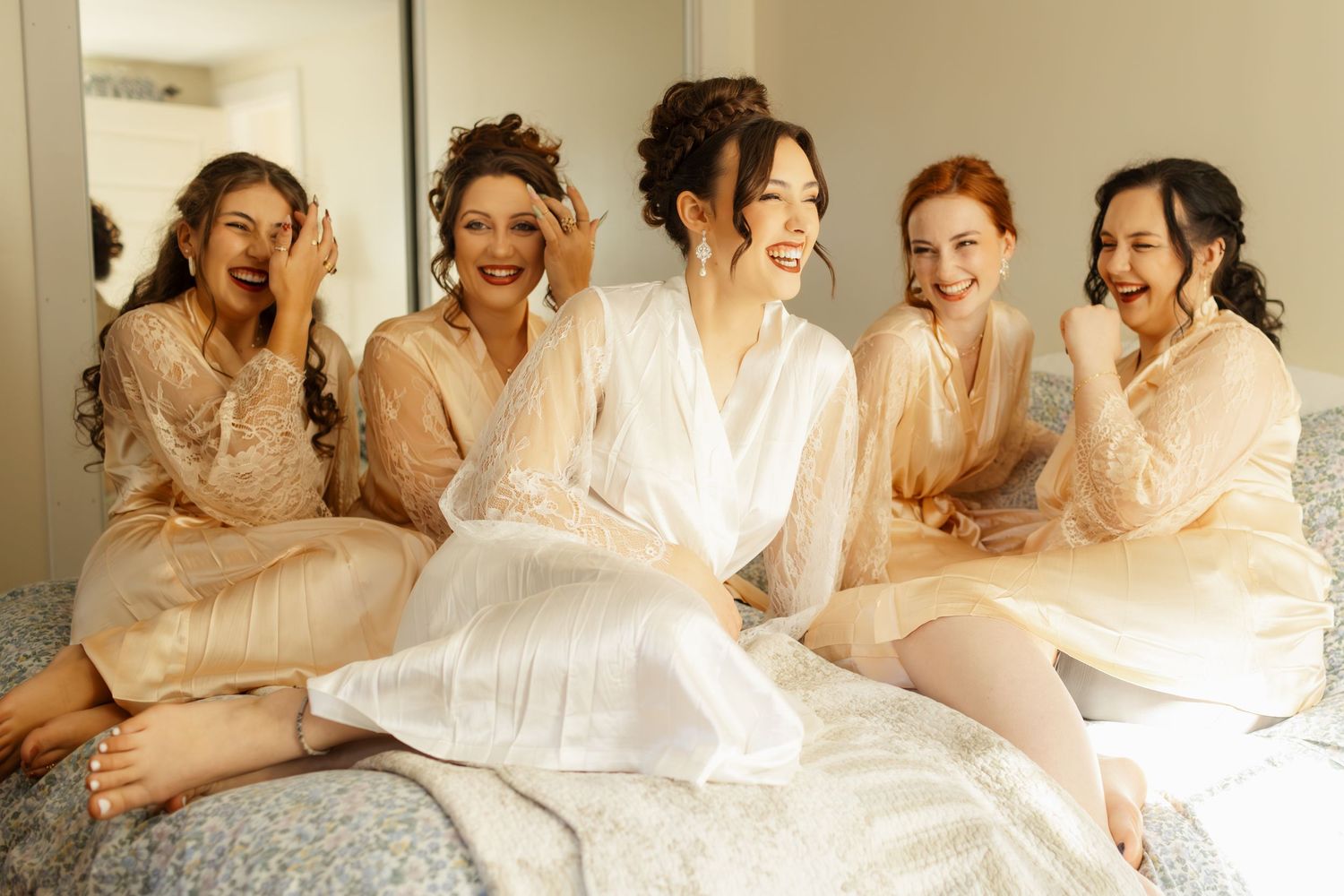 Bride and bridesmaids laughing together in white and cream robes sitting on bed during wedding morning preparations.