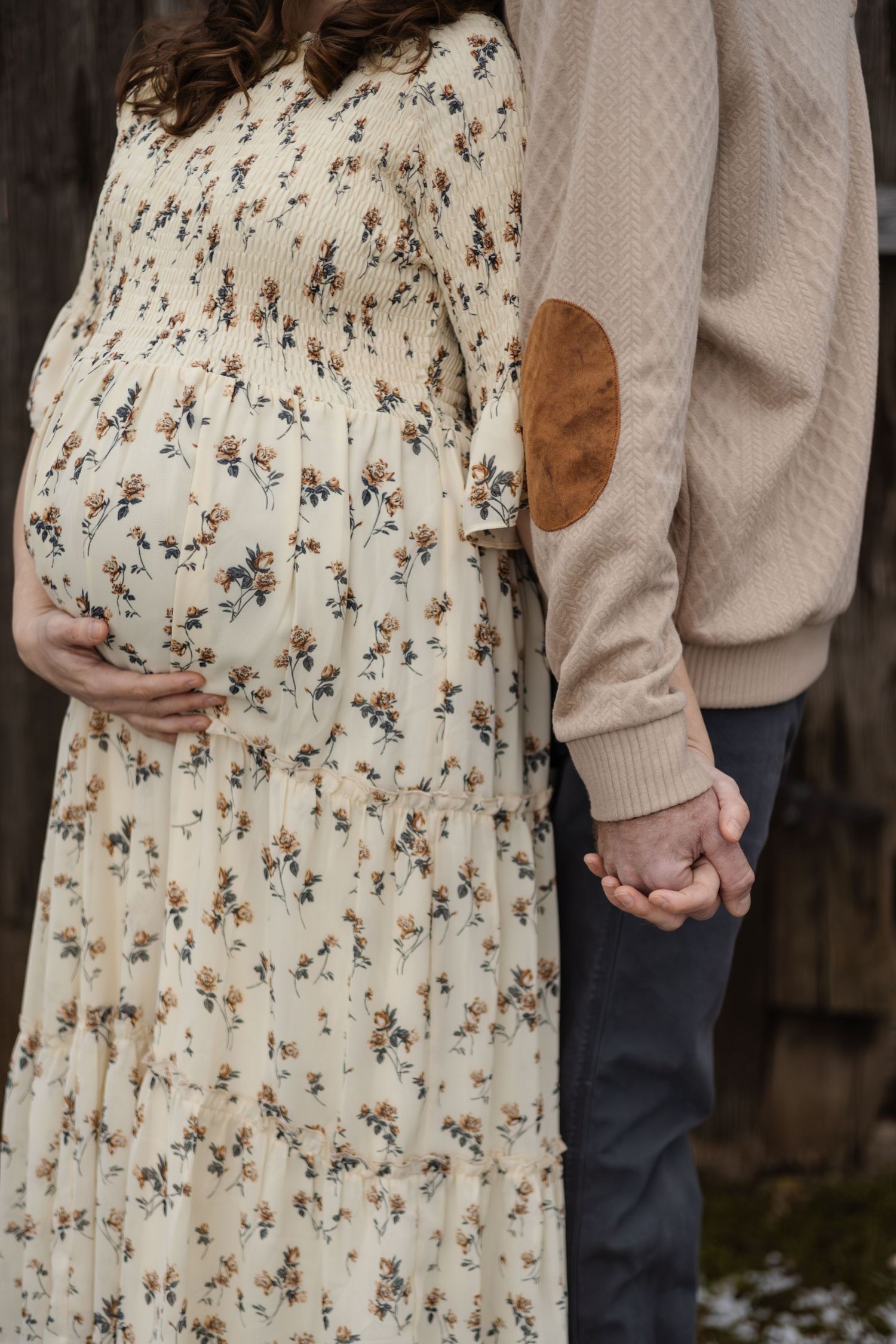 Couple holding hands with floral maternity dress against rustic backdrop.