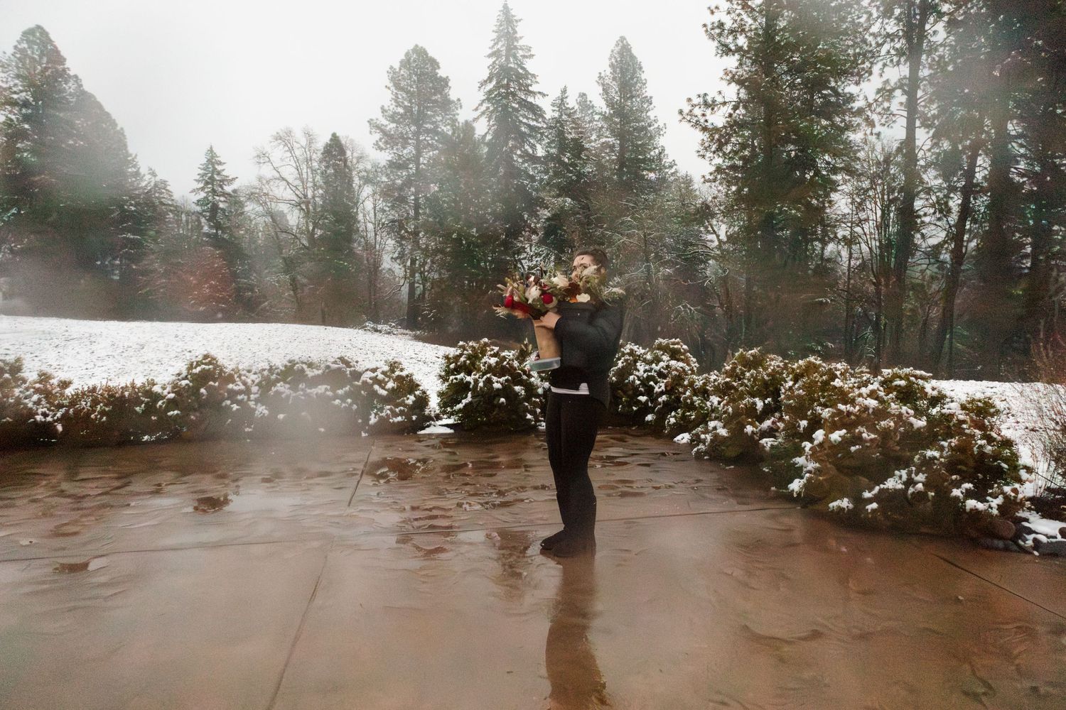Groom holds bride in snowy outdoor setting, surrounded by pine trees and string lights.