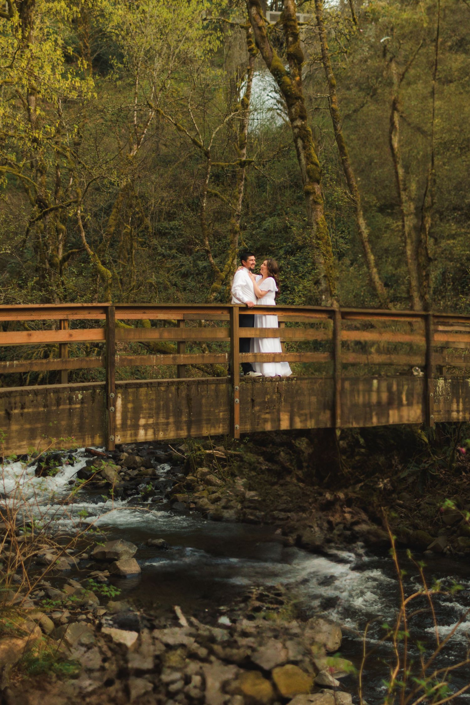 Couple in white embracing on a wooden bridge over a rocky stream surrounded by lush green forest trees.