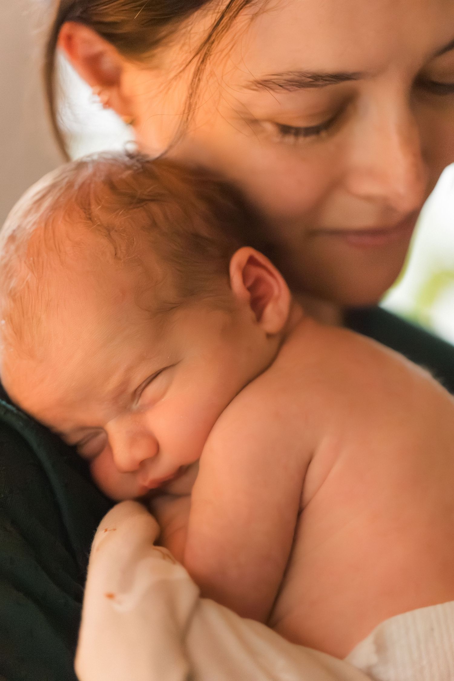 A mother tenderly holds her sleeping newborn baby close to her chest, smiling softly in a warm, peaceful moment.