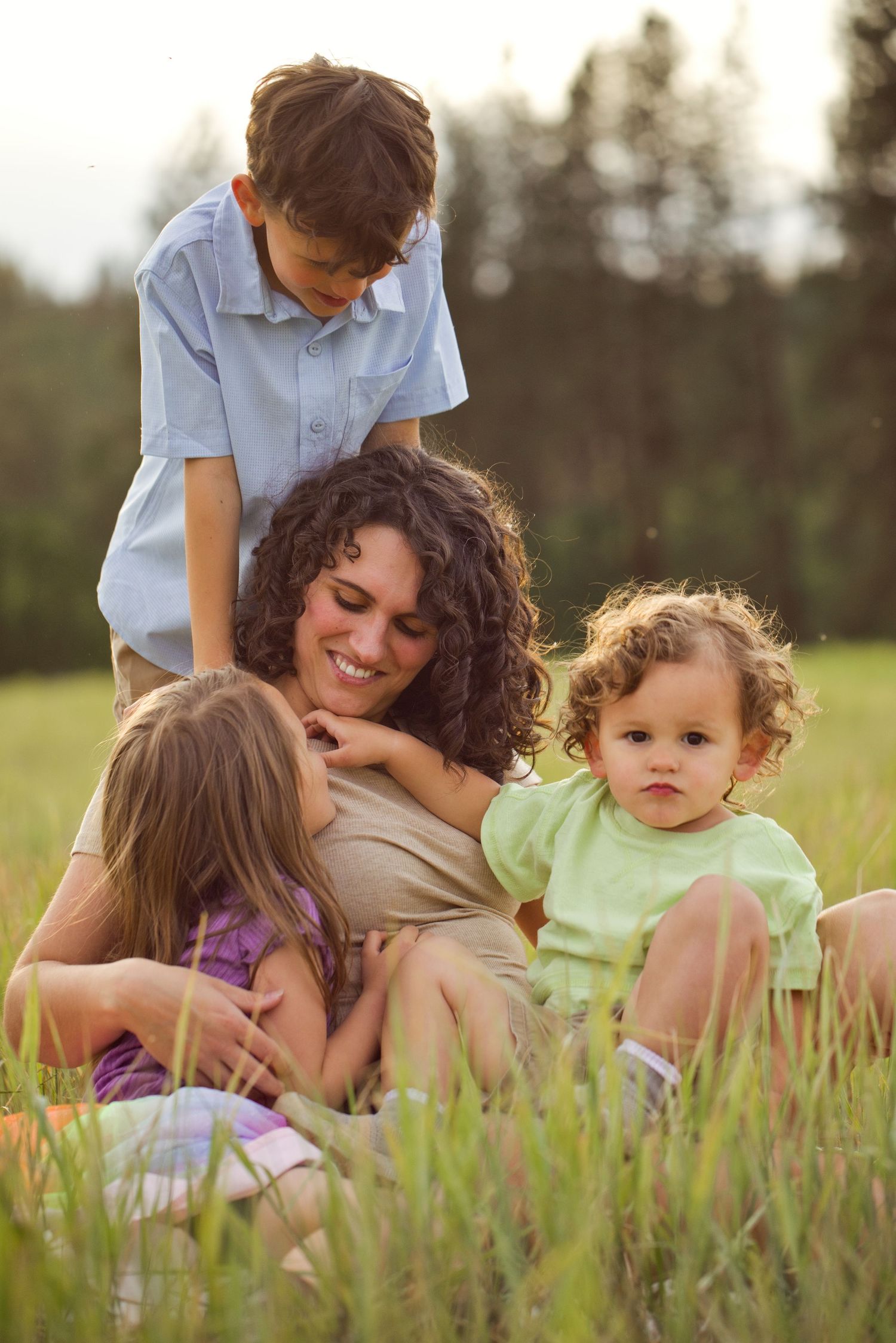 A smiling mother relaxes in a green field with her three young children on a sunny day.