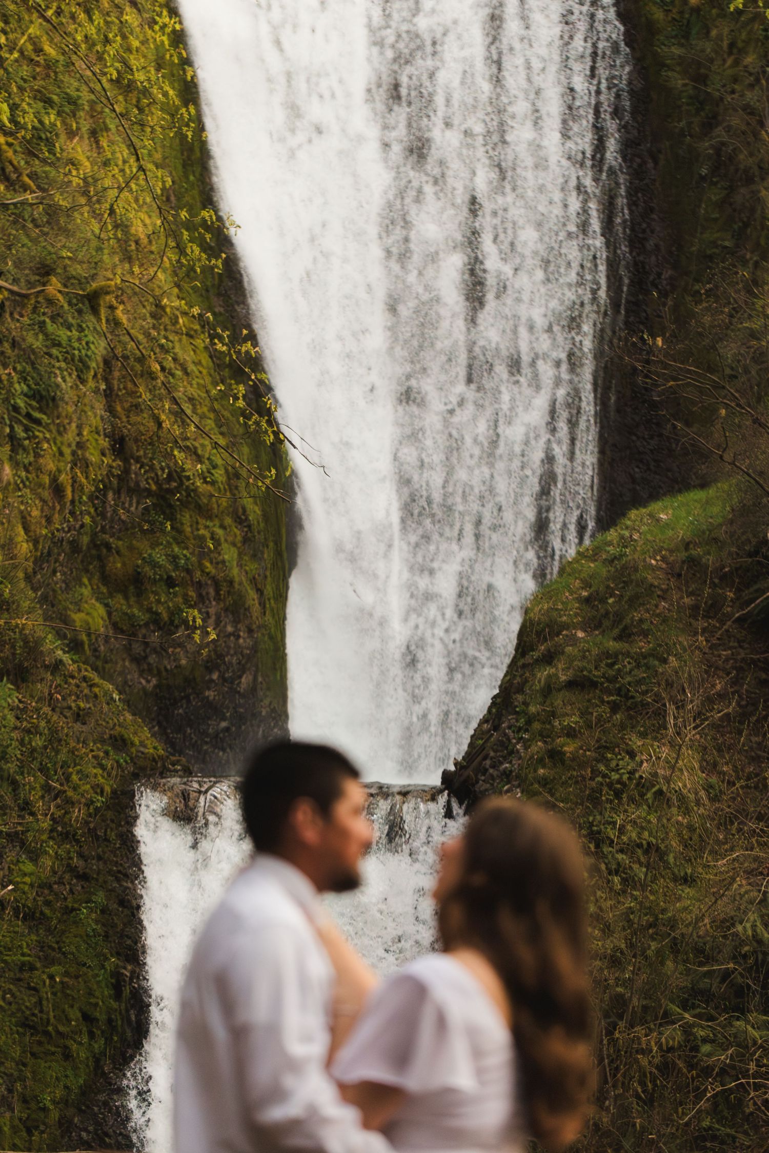 Couple in white attire standing before a majestic waterfall surrounded by lush green moss-covered cliffs.