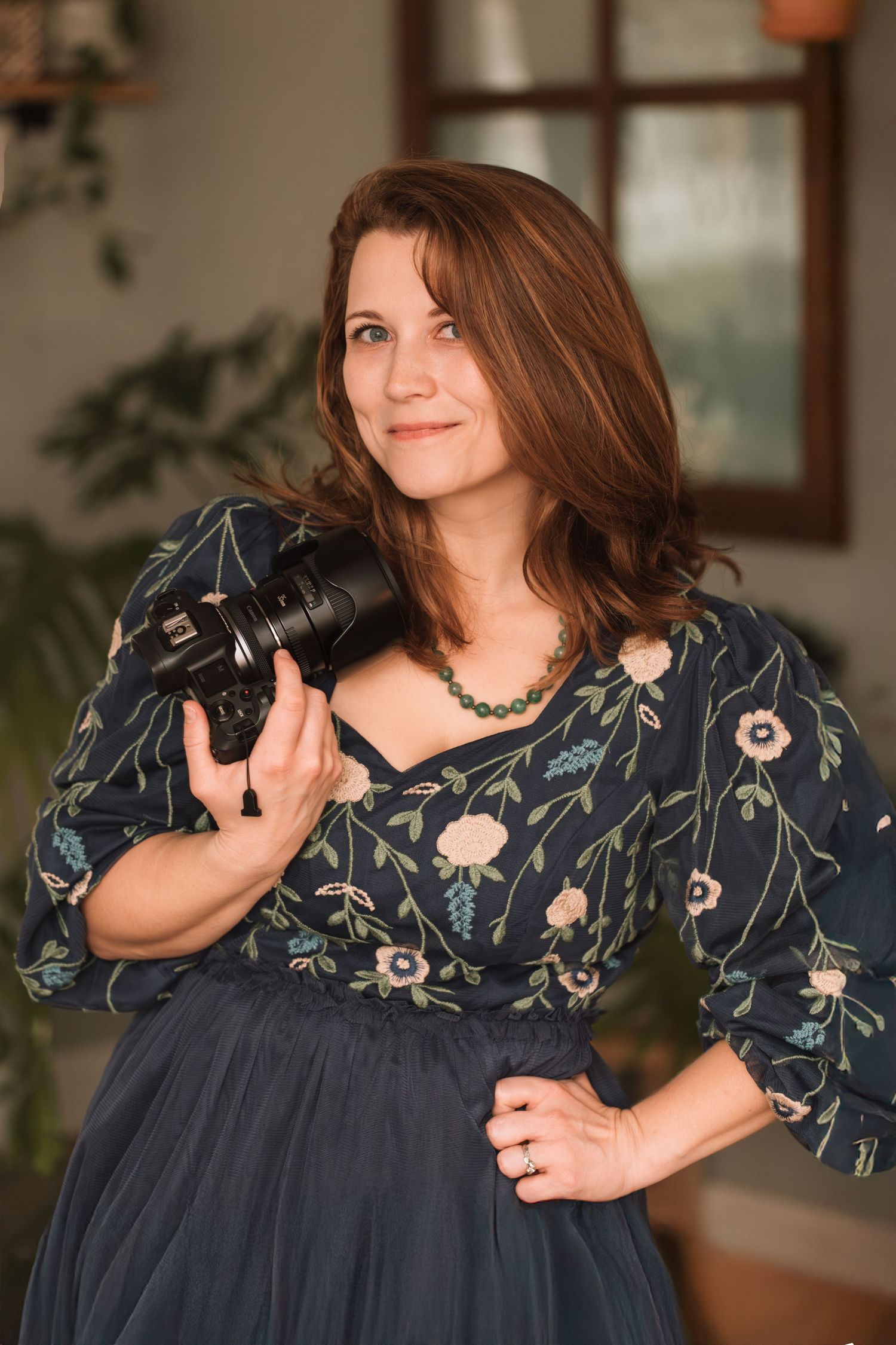 Photographer in floral dress poses with professional camera equipment in indoor setting.