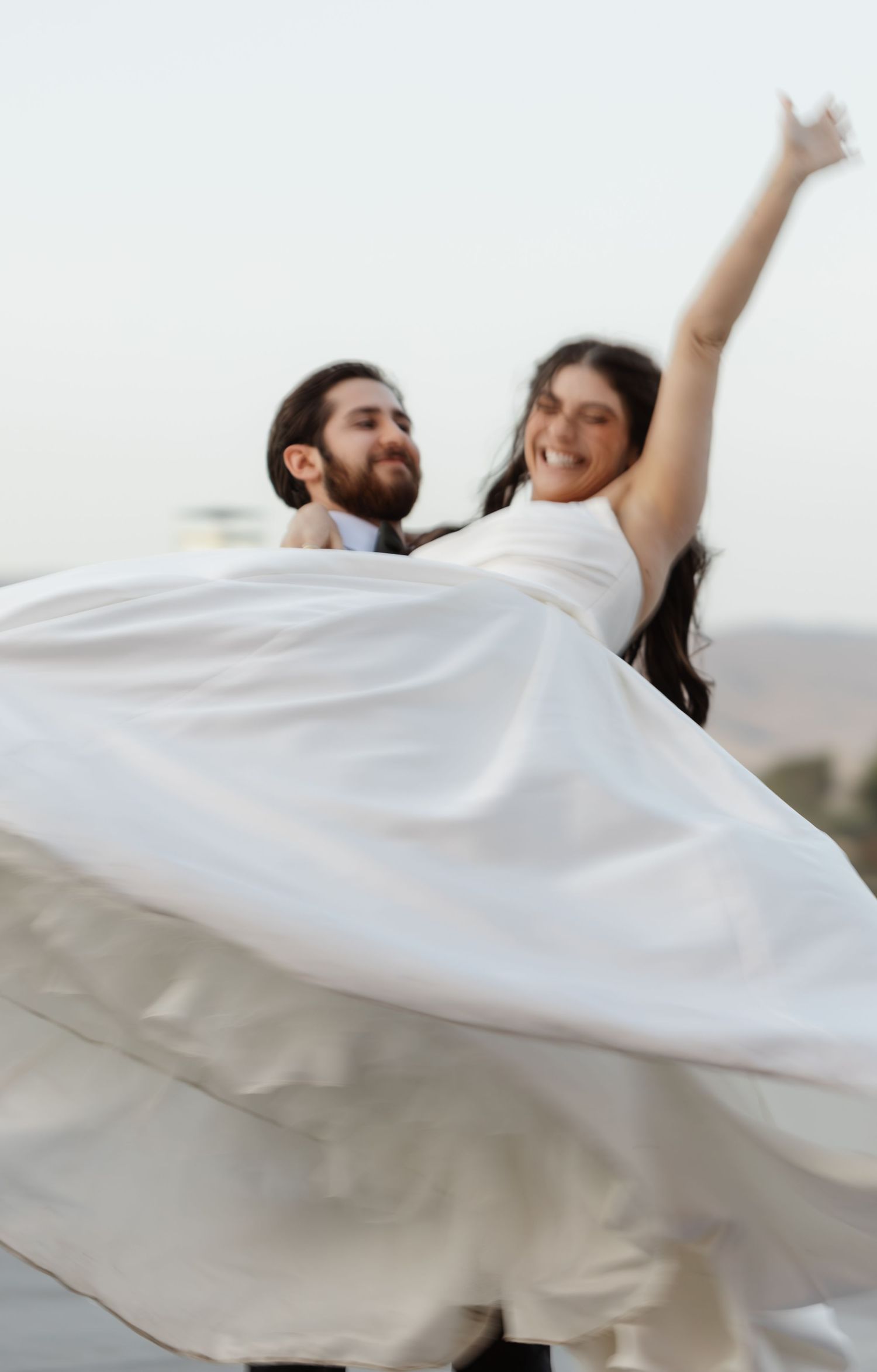 Joyful wedding couple dancing and twirling in white dress against light background.