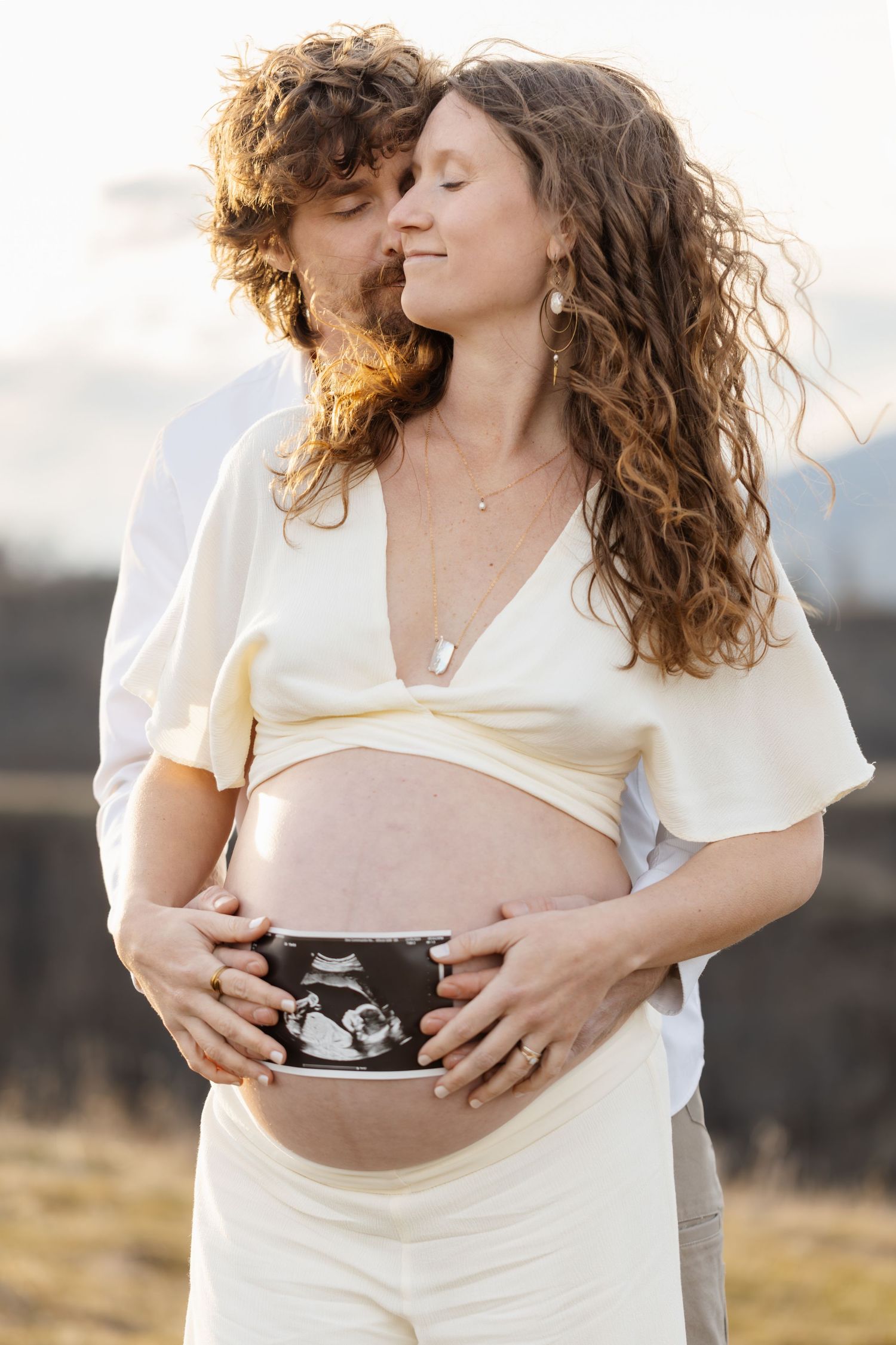 Couple sharing intimate moment outdoors while holding ultrasound photos against sunset.