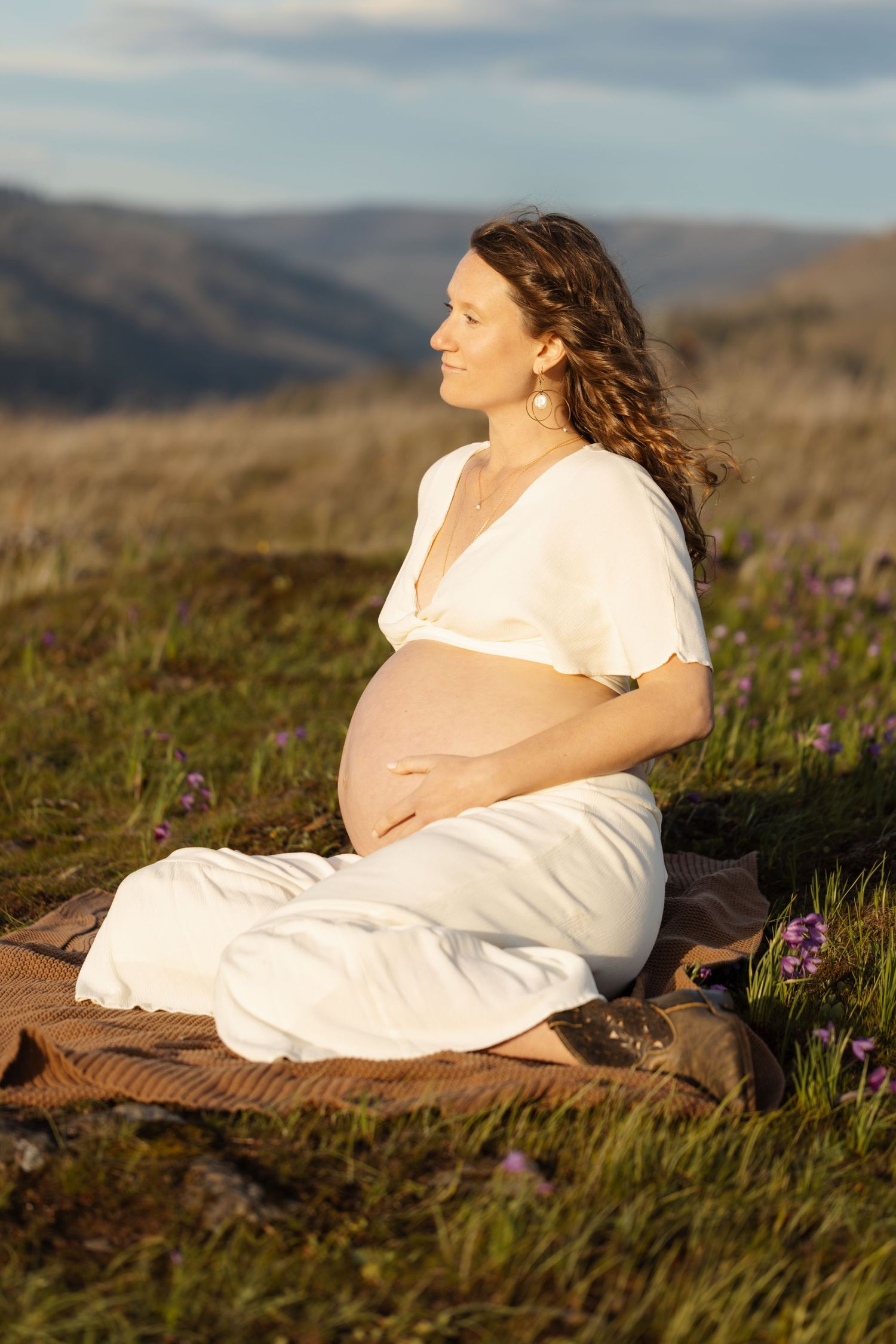 Expectant mother in white dress sitting peacefully in mountain wildflower field at sunset.