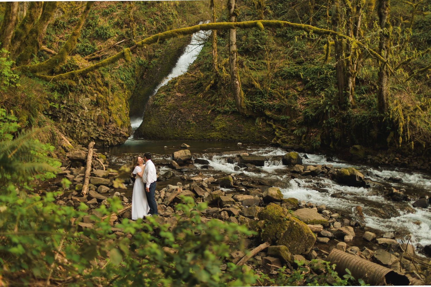 Couple embracing on rocky riverbank with mossy waterfall and lush green forest backdrop.