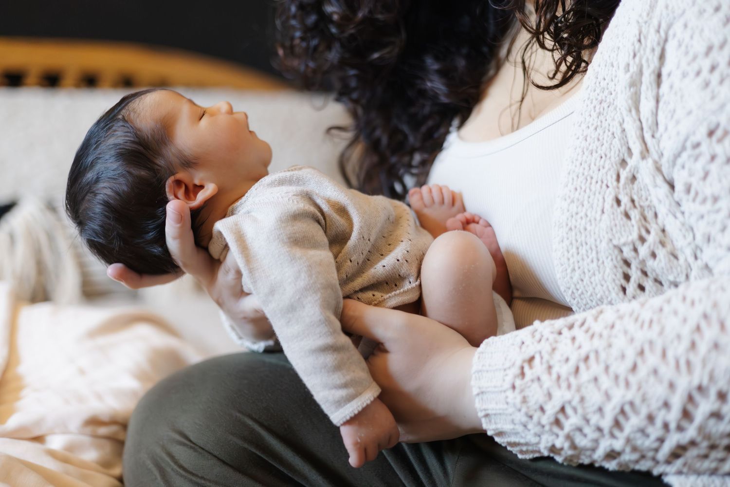 A baby in a cream sweater lies cradled in someone's lap wearing white lace.