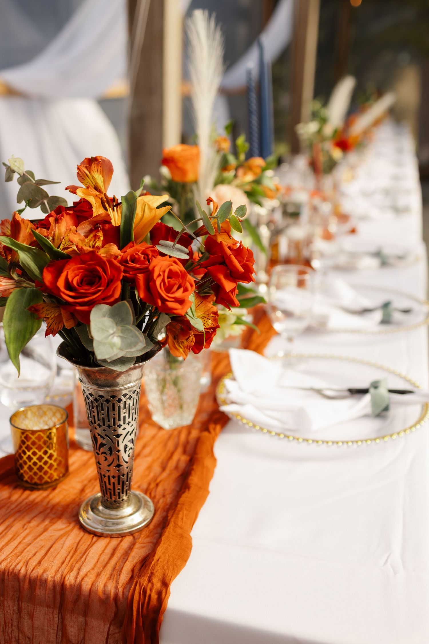 Elegant wedding table setting with orange roses in silver vase and burnt orange runner on white tablecloth.