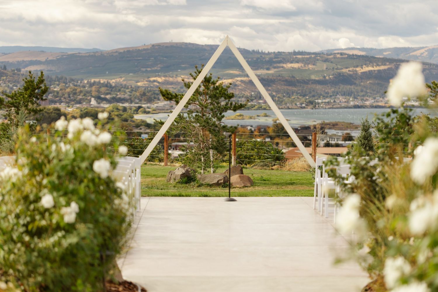 A panoramic view of a scenic wedding ceremony setup with white triangular arches overlooking mountains and a lake.