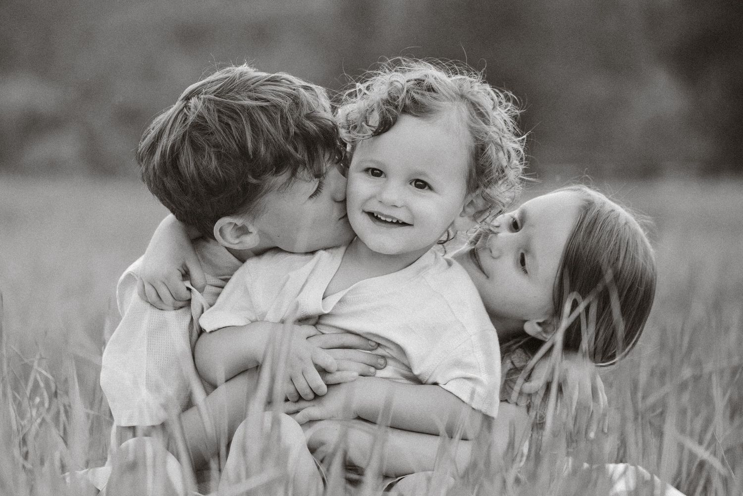Black and white photo of two children kissing a smiling toddler in a grassy field.