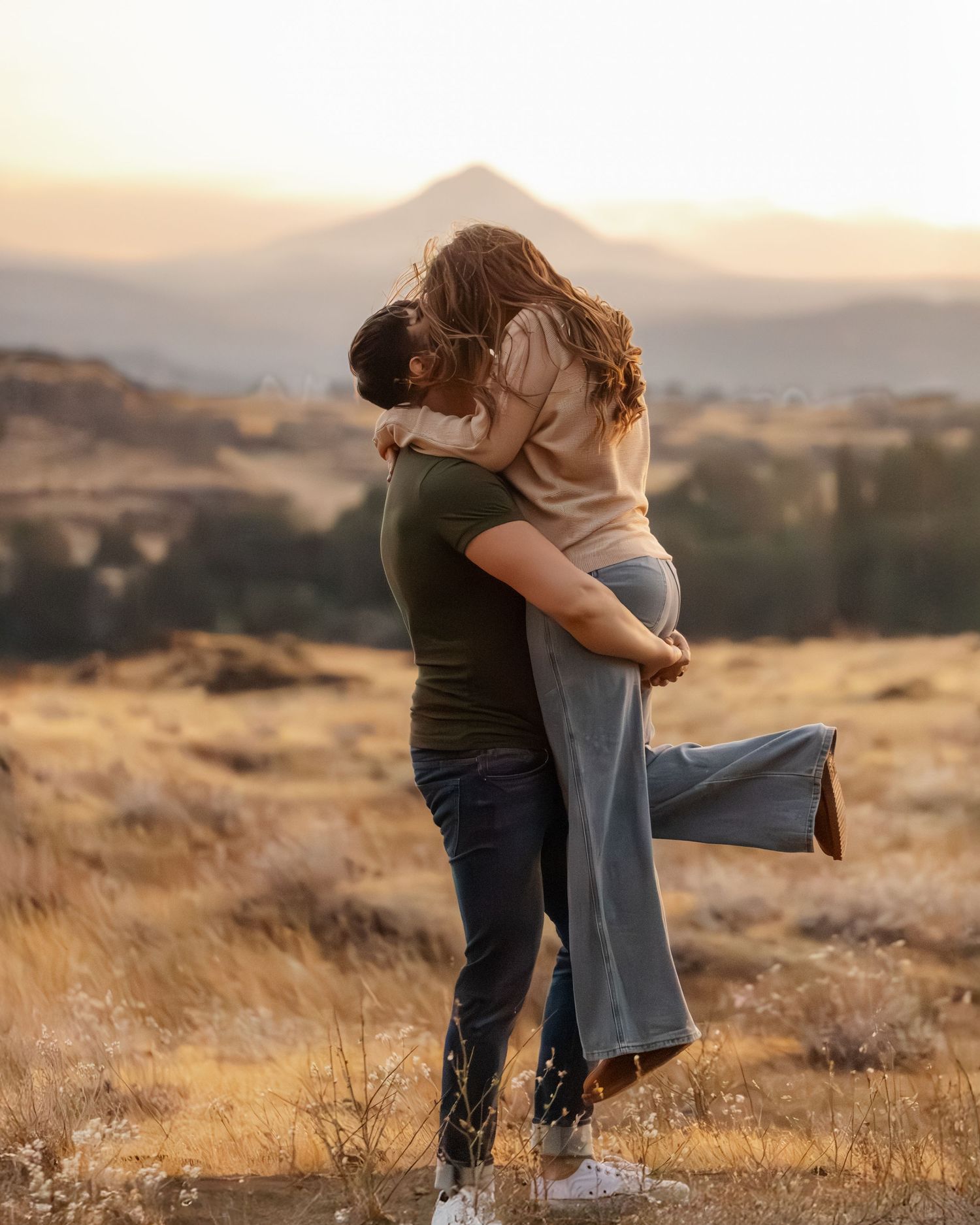 A romantic couple embraces in a golden field at sunset with mountains in the background.