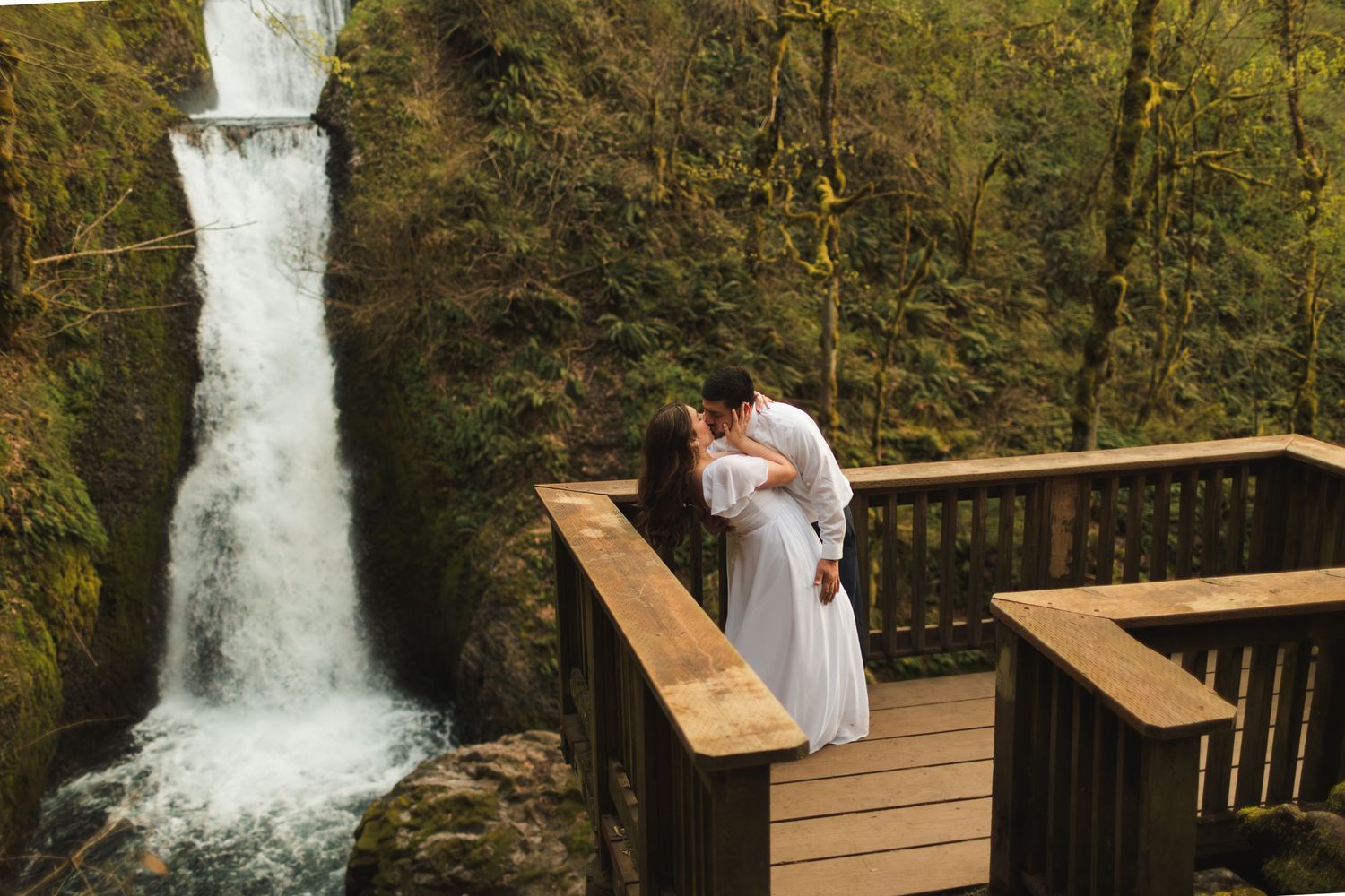 Couple sharing a romantic kiss on a wooden deck overlooking a stunning waterfall surrounded by lush green moss.