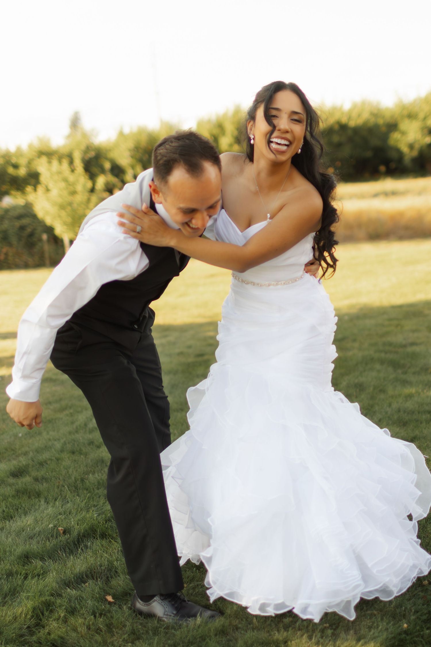 Laughing bride in a white ruffled strapless wedding gown hugs a groom in a black vest and slacks on a sunny grassy field.