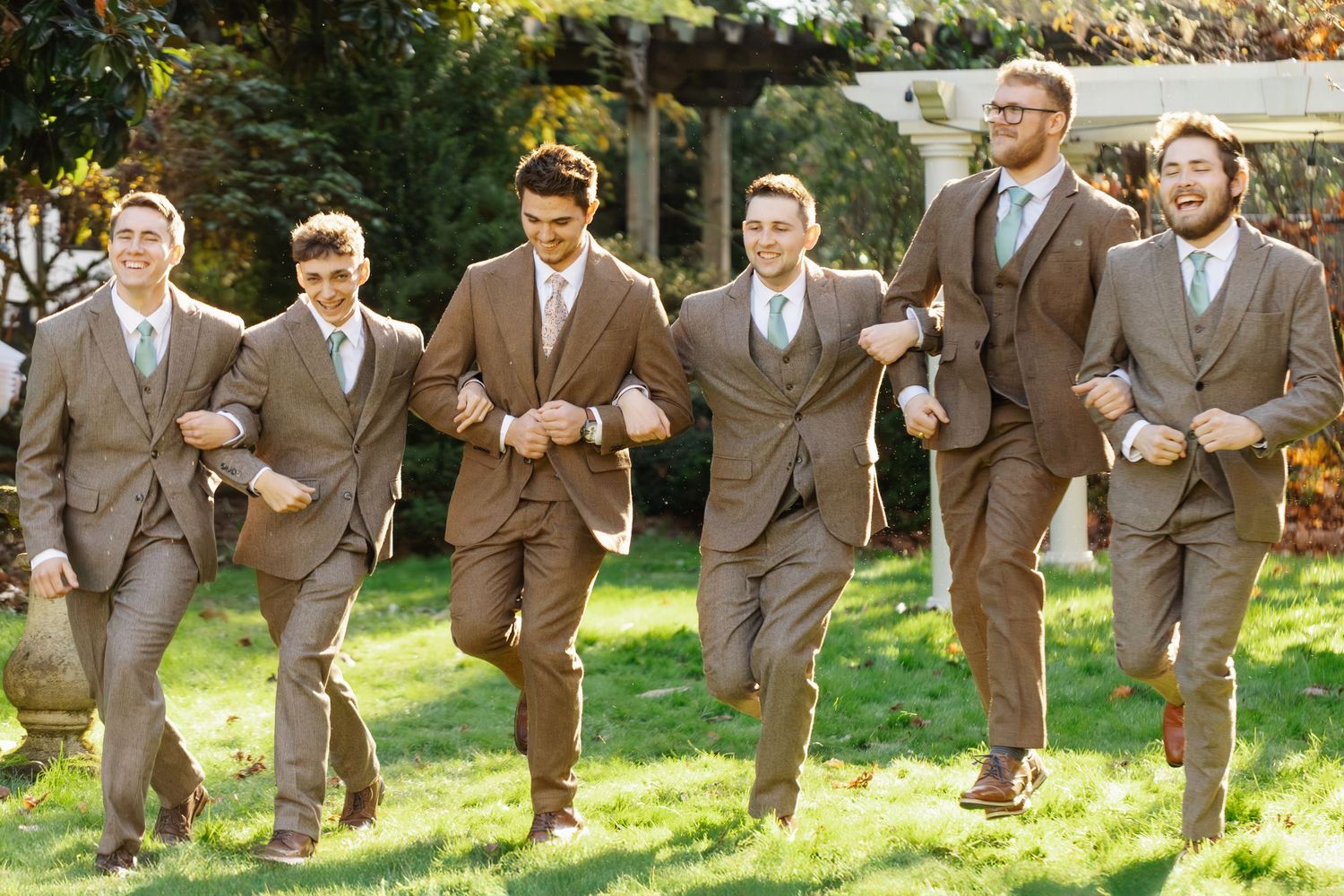 Groomsmen in matching brown suits running and laughing together on a sunny lawn during a wedding celebration.