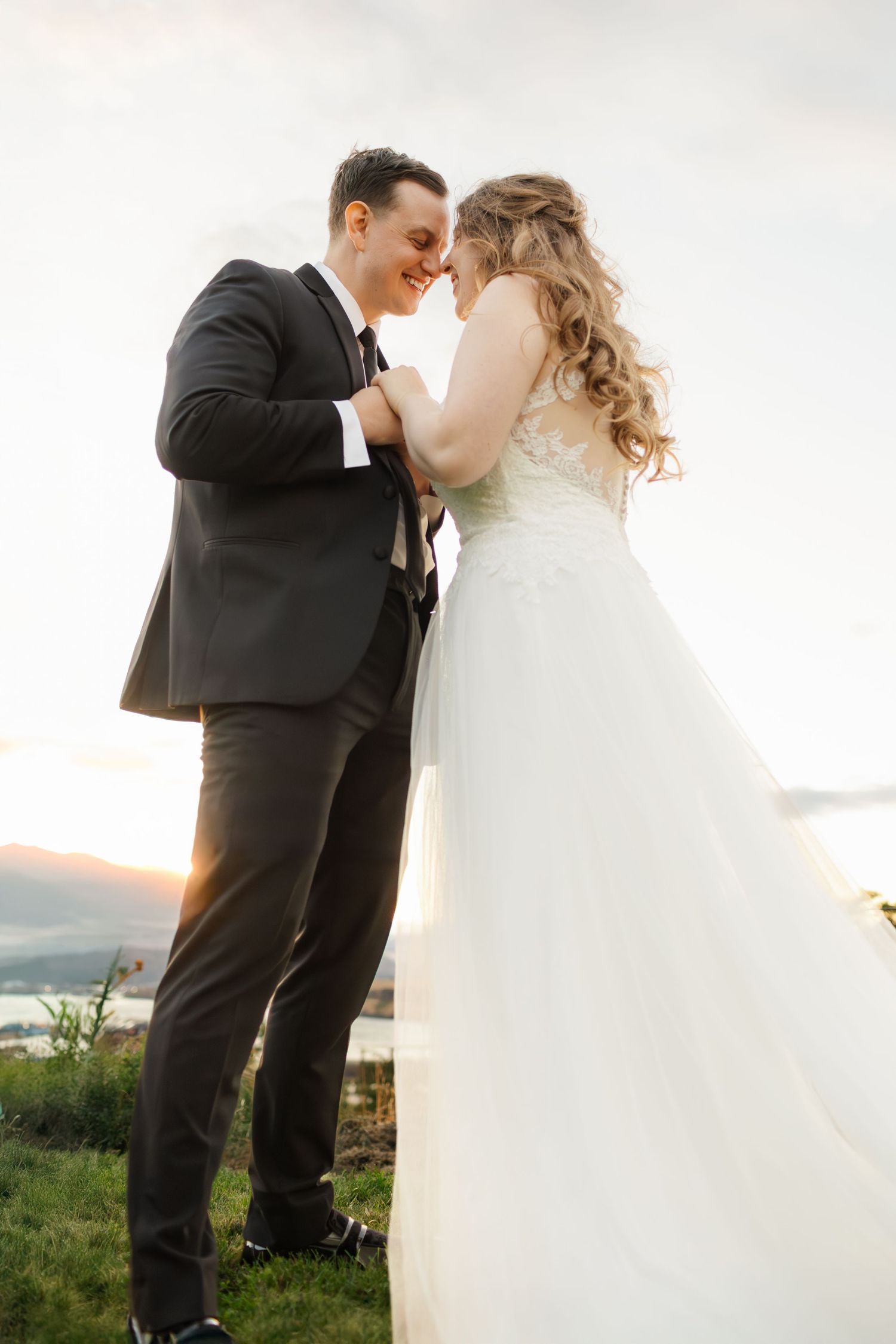 A romantic wedding couple embraces outdoors at sunset with mountains in the background.