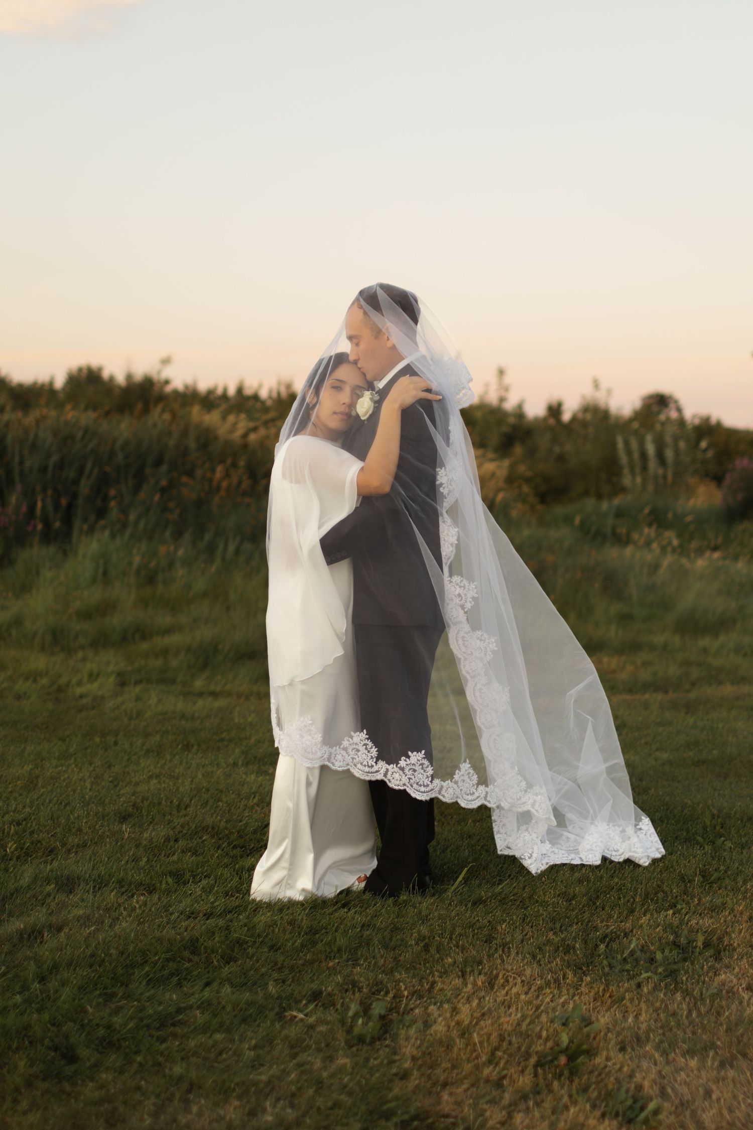 A romantic wedding couple embraces in a field at sunset with a flowing white veil creating a dreamy silhouette.