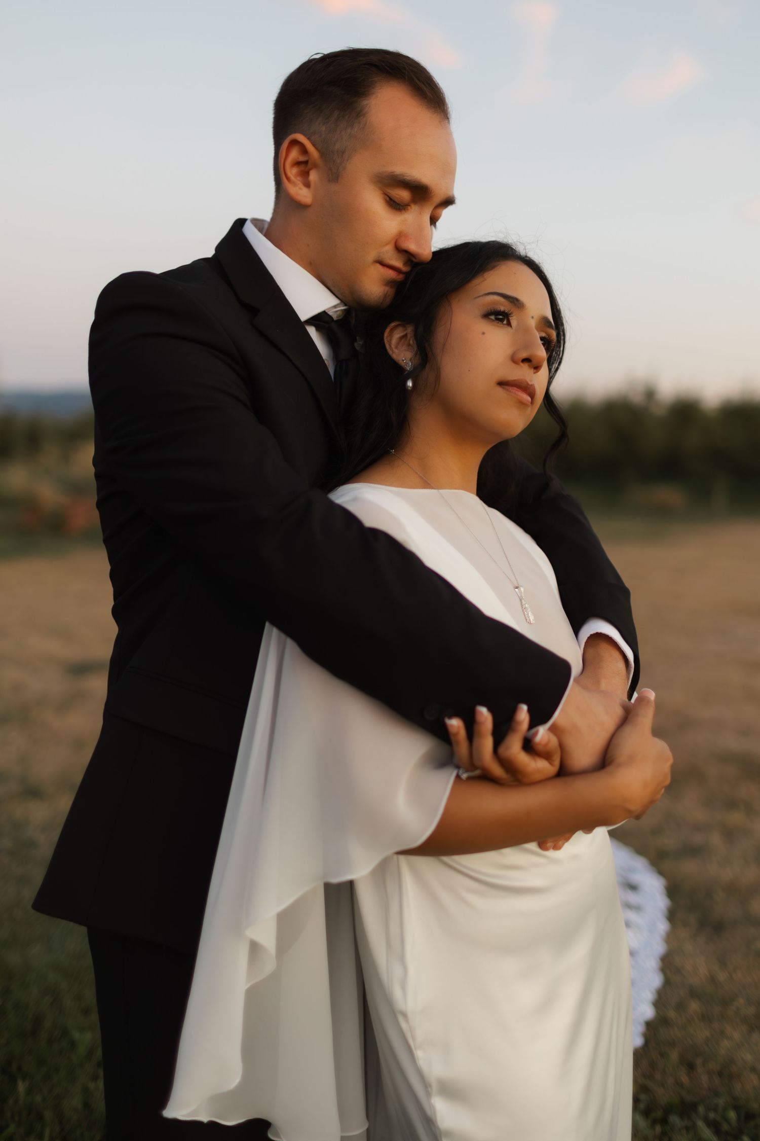 A romantic sunset wedding portrait shows a couple embracing in formal attire against a scenic outdoor backdrop.