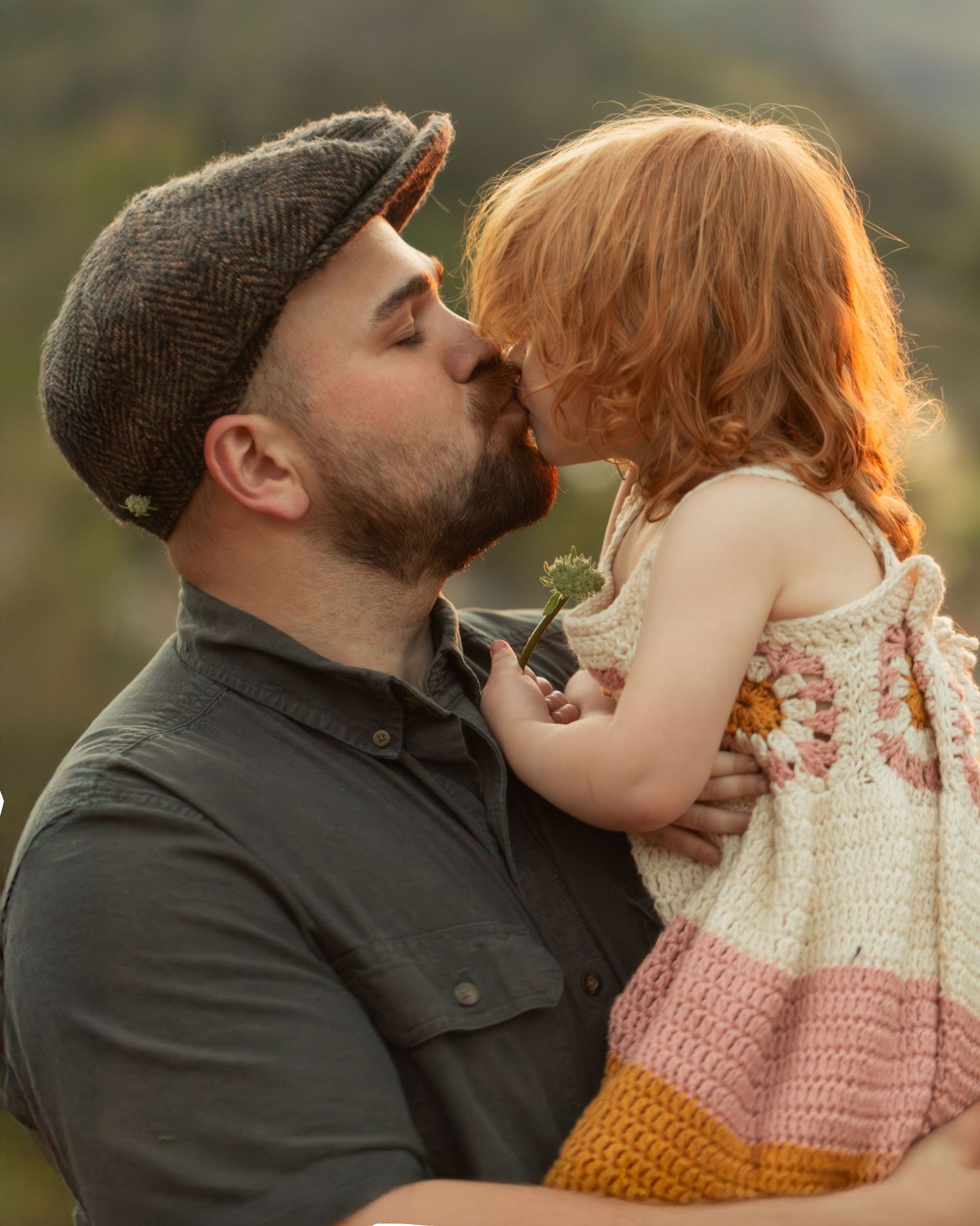 A bearded man in a flat cap kisses a red-haired toddler girl in a crochet dress outdoors.