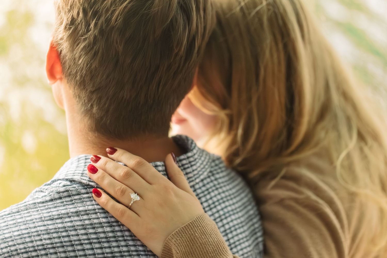 Woman with red manicured nails and a diamond engagement ring wraps her arm around a man in a plaid jacket in a warm embrace.