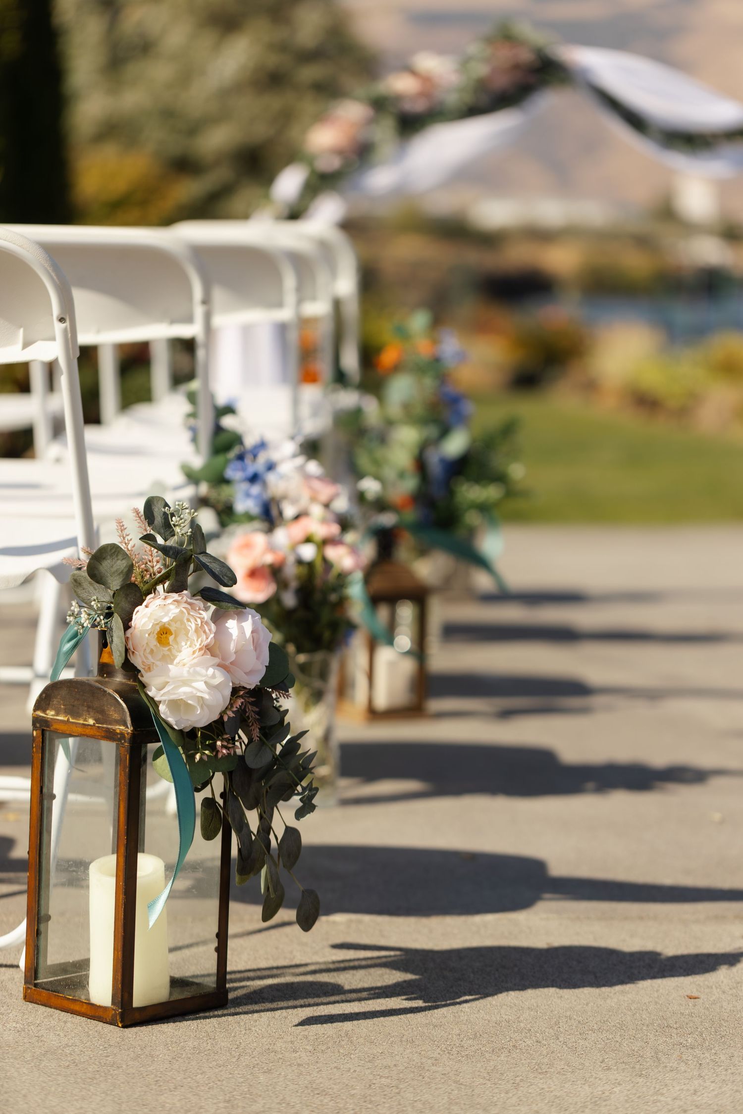 Elegant wedding aisle lanterns with white candles and blush floral arrangements beside white chairs at an outdoor ceremony.