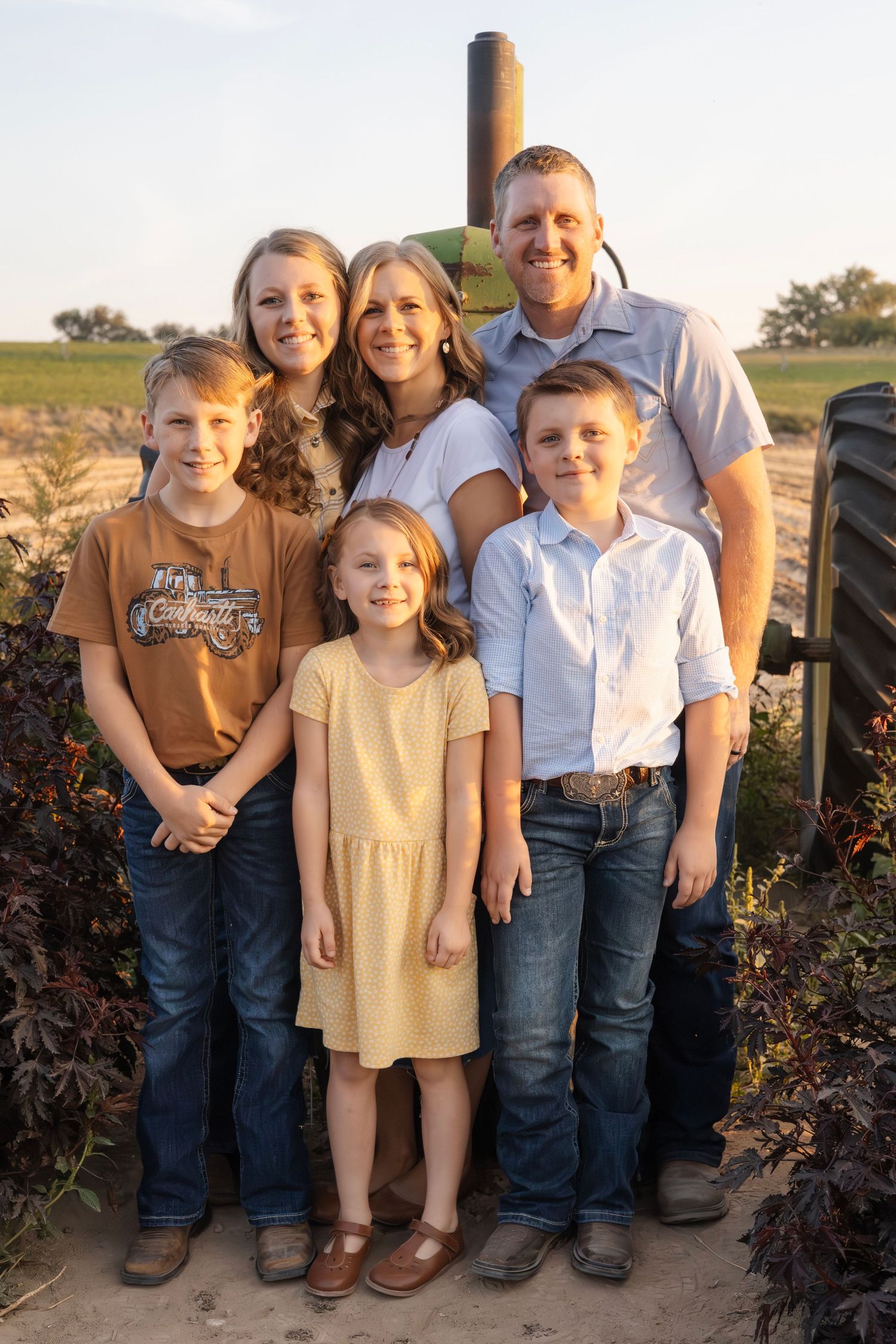 Family poses together in casual attire against rustic farm equipment backdrop.