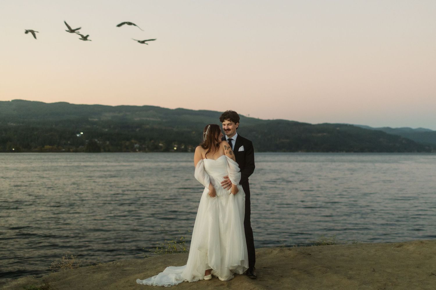 Bride and groom embrace along the Columbia River at The Locks in Cascade Locks, Oregon, as geese fly overhead at sunset.