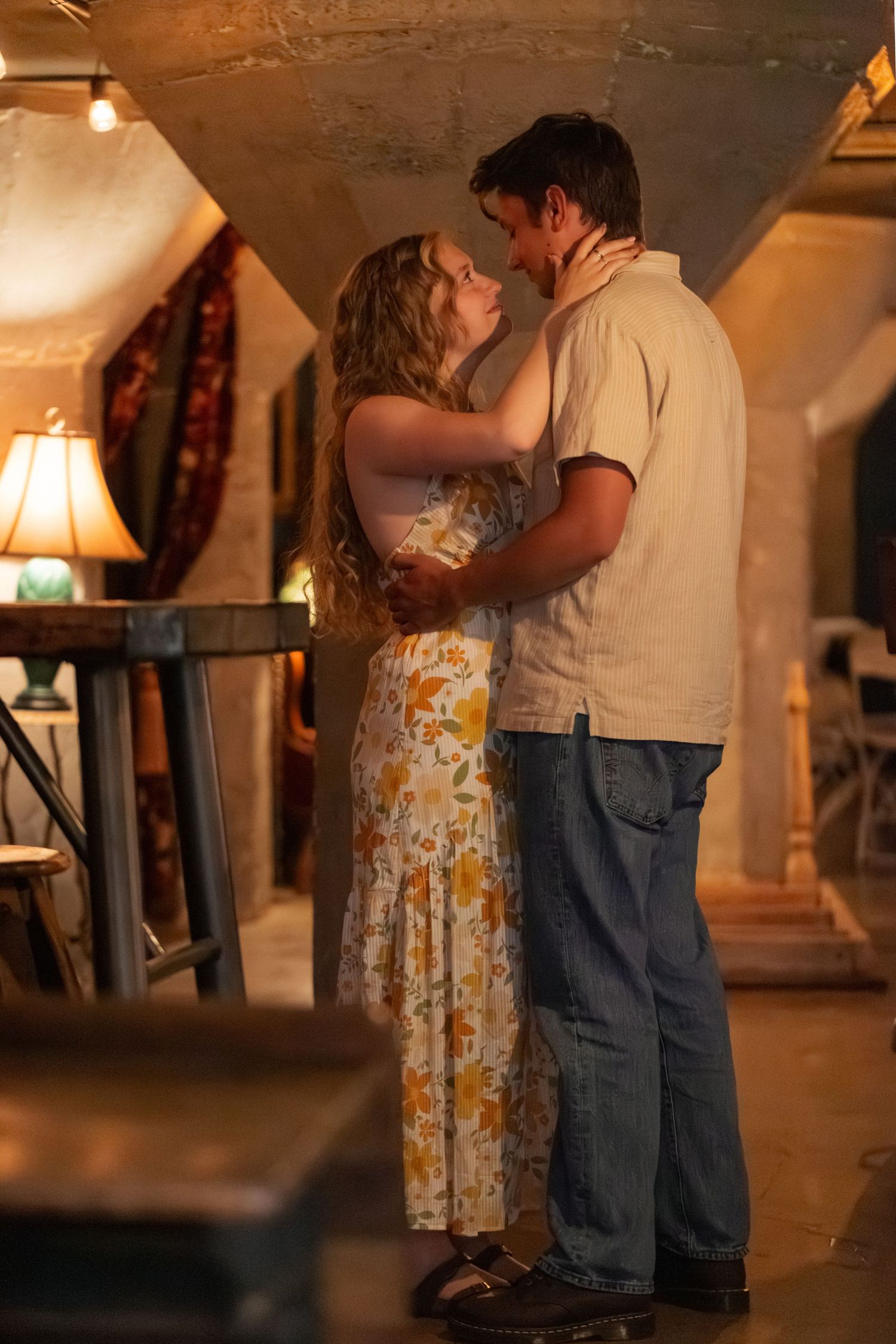 A young couple in a romantic embrace, woman in floral dress and man in jeans, sharing an intimate moment indoors.