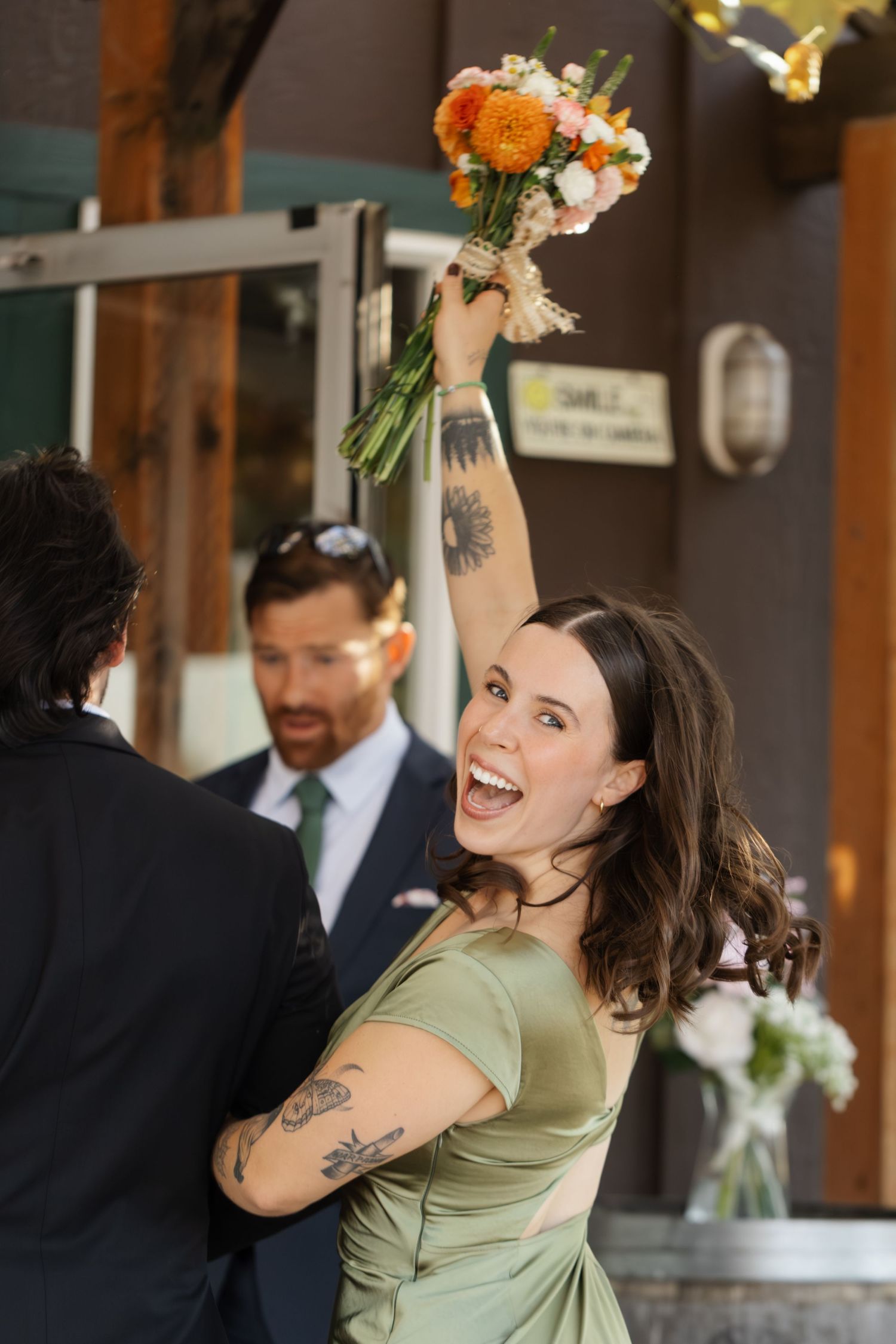 A bridesmaid in a sage green dress joyfully dances while holding a vibrant orange and white bouquet.
