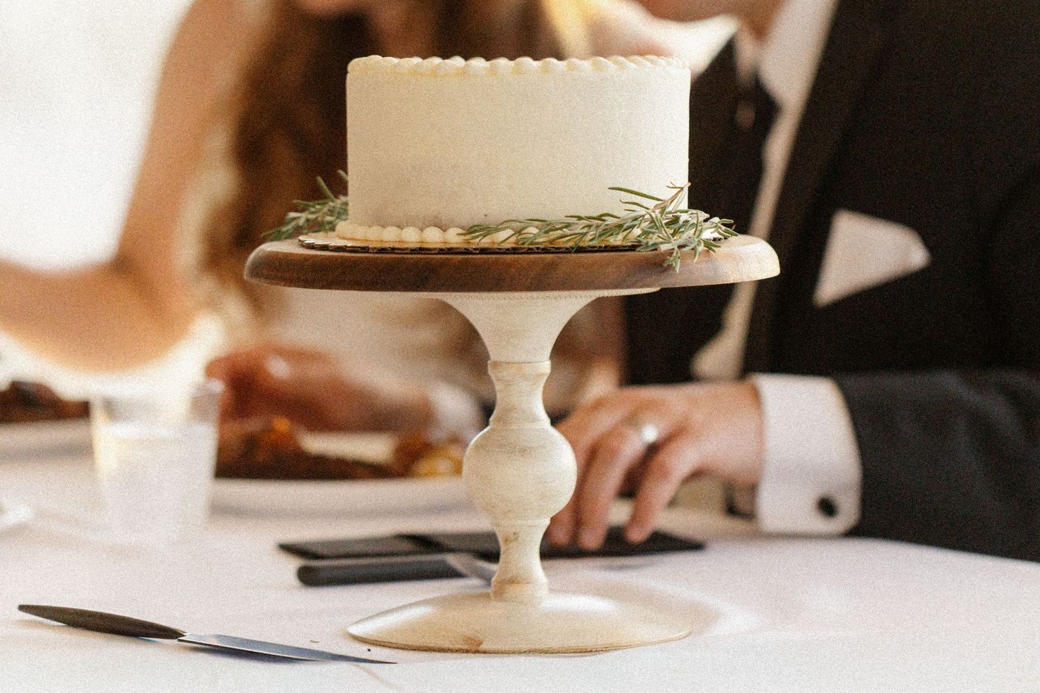 Simple white wedding cake displayed on an elegant pedestal stand against a blurred background.
