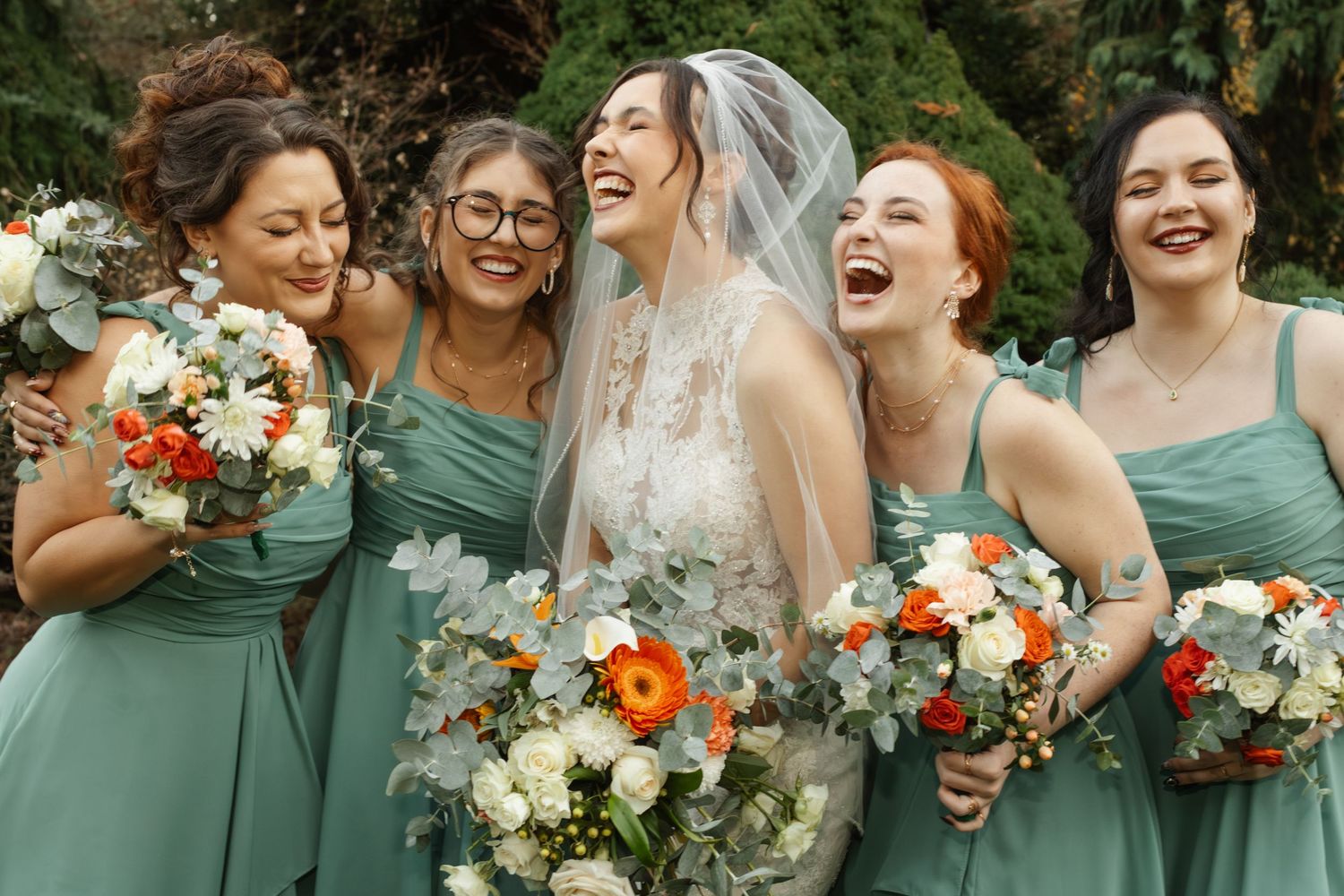 A bridal party in sage green dresses laughs together while holding vibrant coral and white floral bouquets.