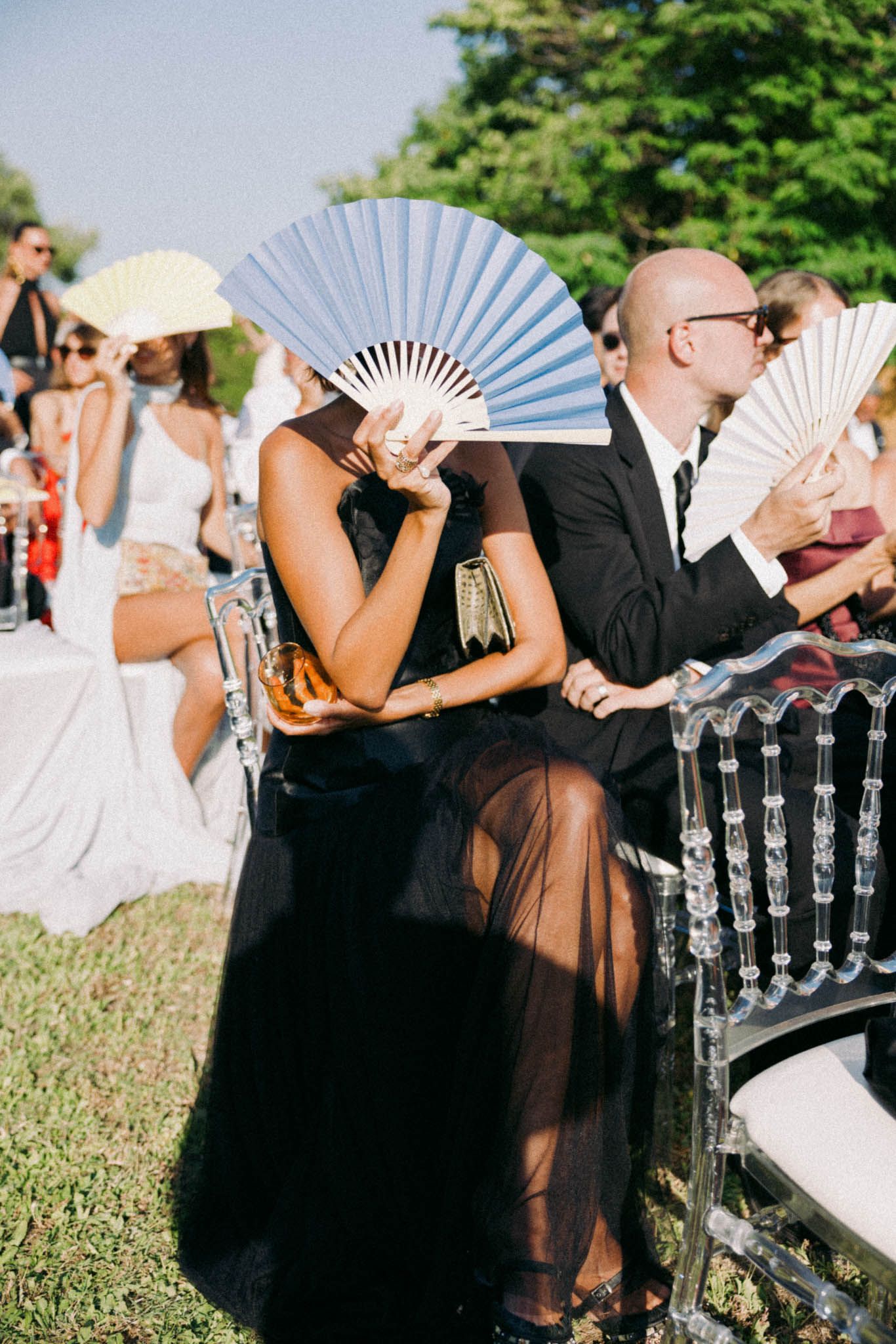 Wedding guests hold white paper fans in front of their faces while seated in black chairs at an outdoor ceremony.