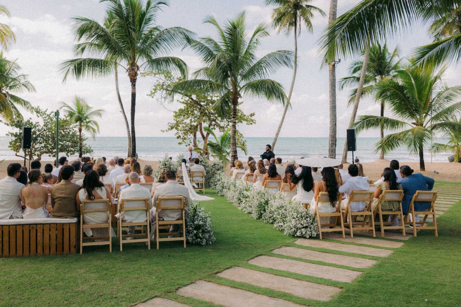 A tropical beach wedding ceremony setup with white chairs and palm trees overlooking the ocean at sunset.