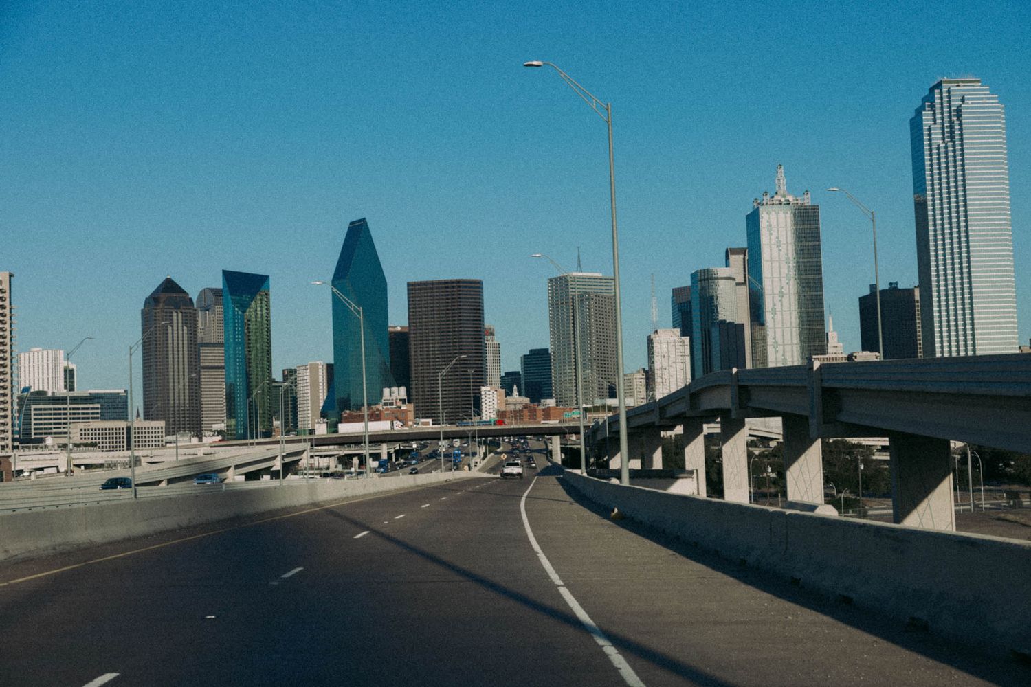 Highway road leading towards downtown Dallas skyline with modern skyscrapers under clear blue sky.