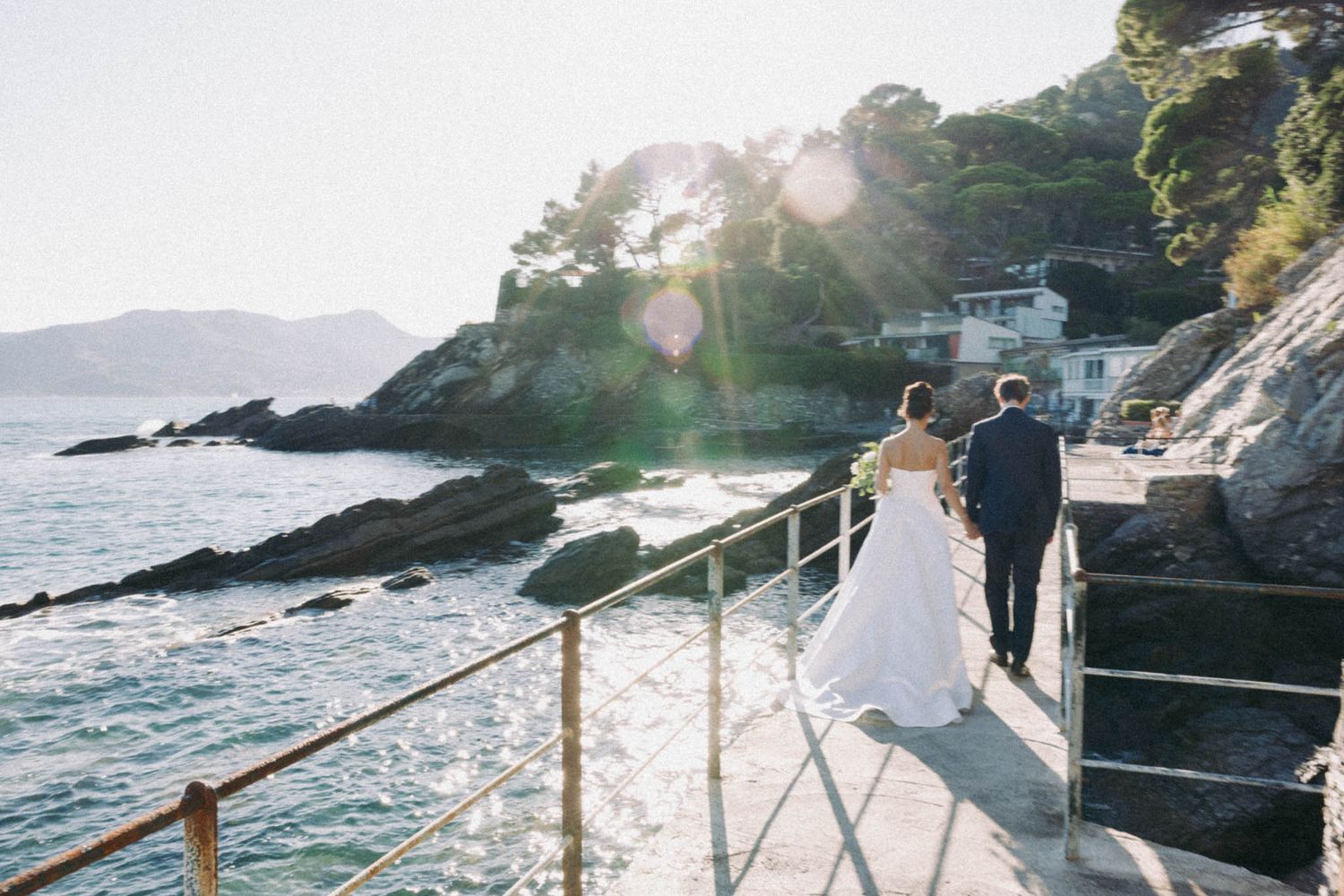 Romantic coastal wedding photoshoot at sunset with couple walking along rocky cliffs by the ocean.