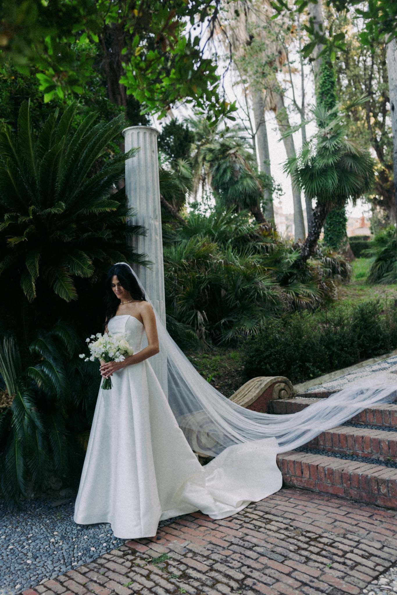 A bride in a stunning white wedding gown with a flowing cathedral veil stands on a brick pathway surrounded by lush greenery.